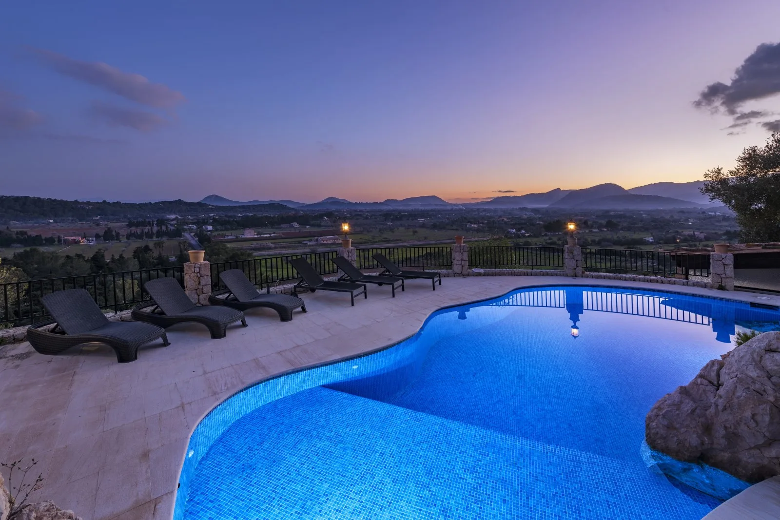 An outdoor swimming pool beside a paved patio overlooks a valley at sunset.