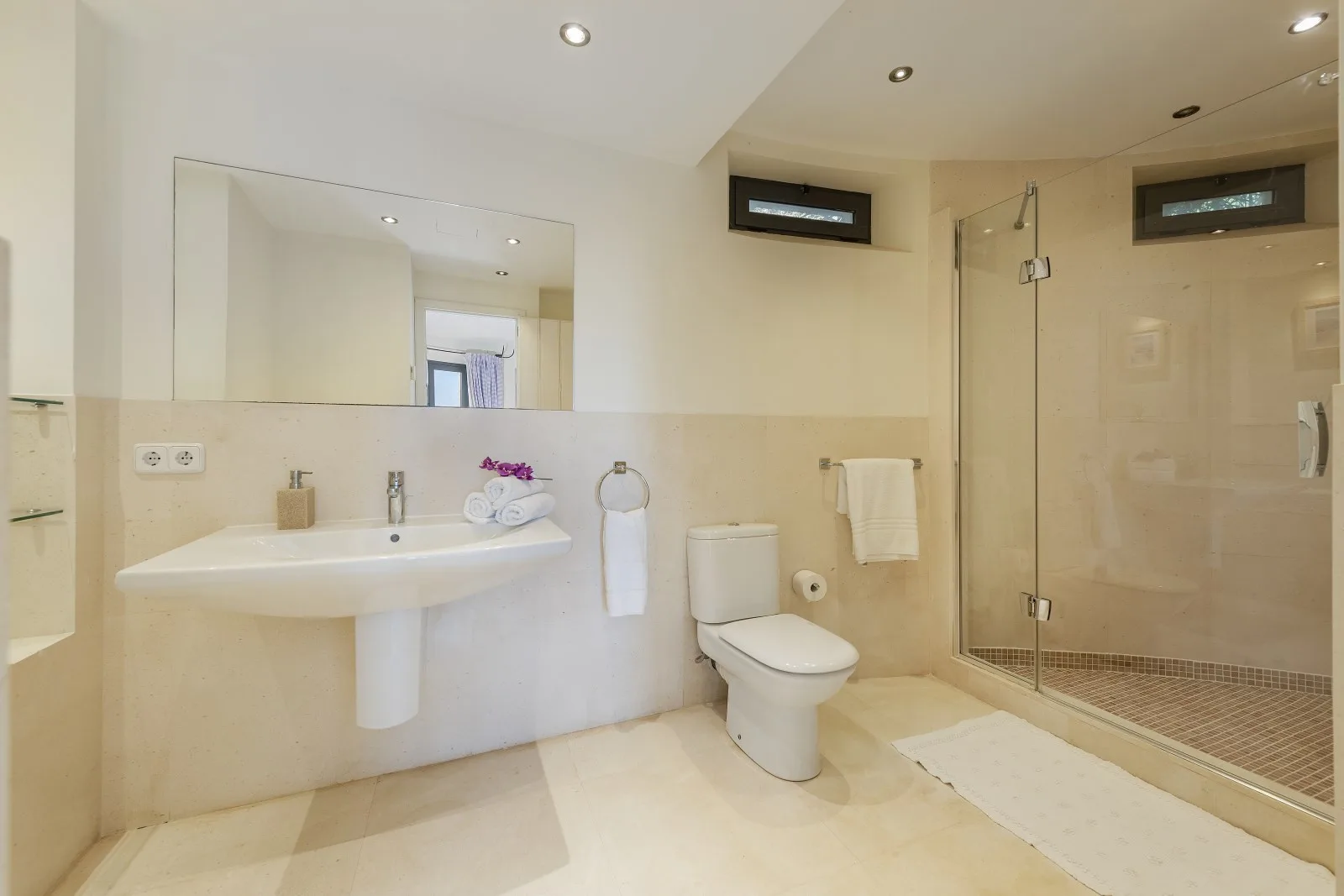 A bright bathroom with a wall-mounted sink, toilet, and glass shower.