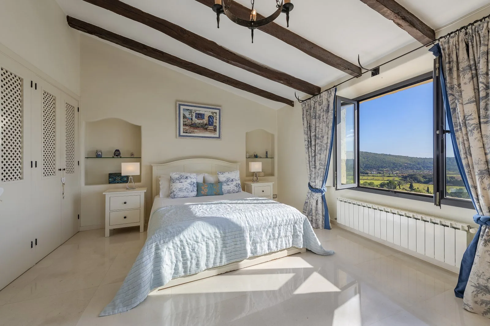 A bright bedroom with a bed beneath dark ceiling beams and a large window.