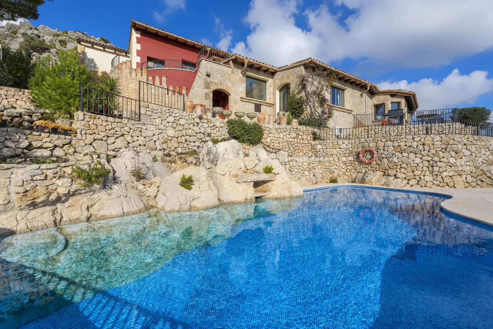 A swimming pool sits below a stone terrace and rustic hillside buildings.
