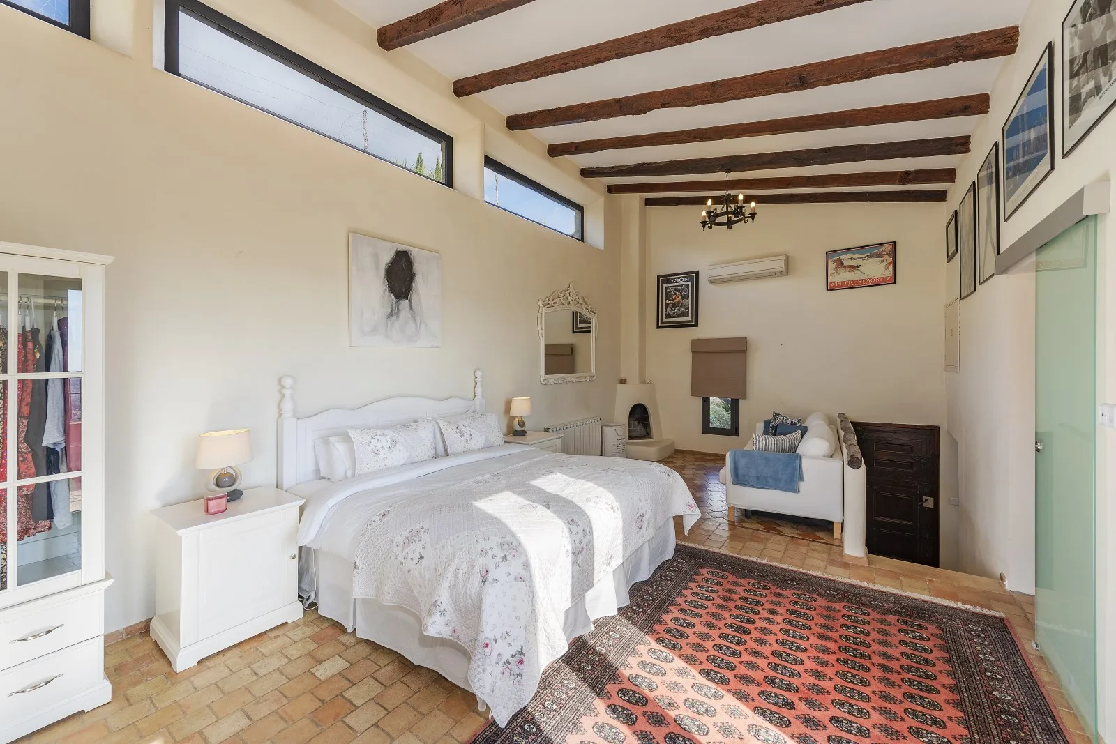 A bright bedroom with a large bed, patterned rug, and exposed ceiling beams.