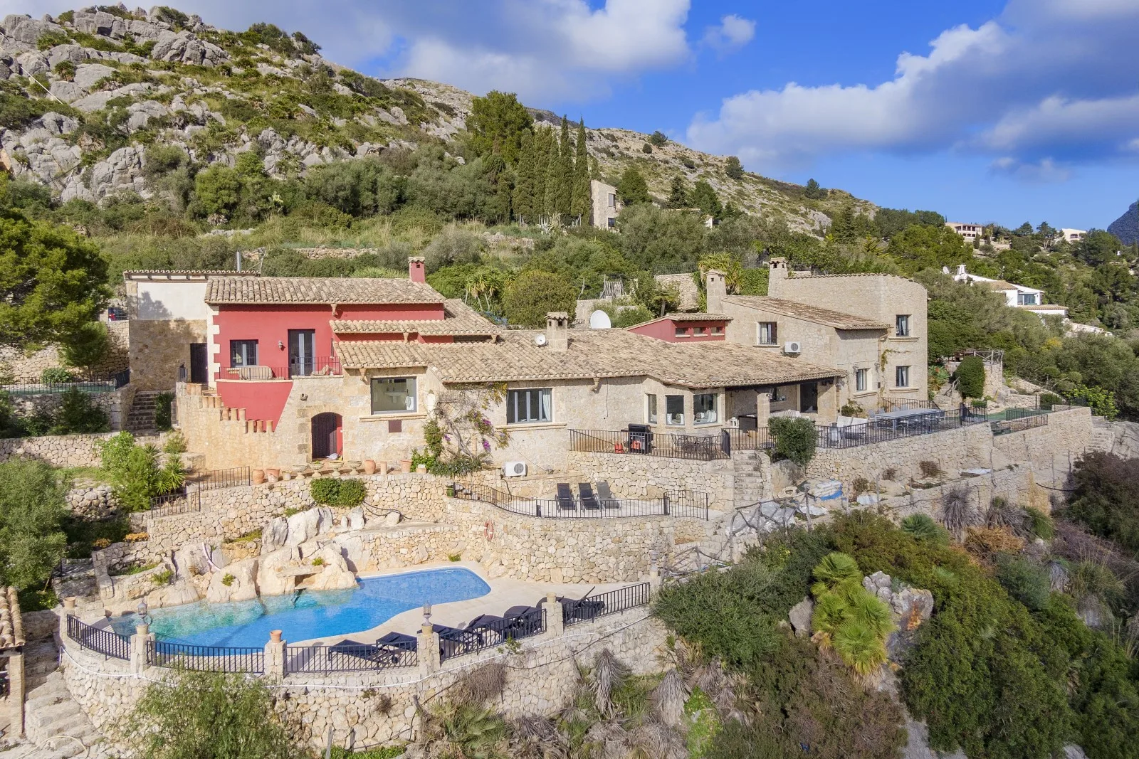 A hillside stone house overlooks a swimming pool and surrounding rocky landscape.