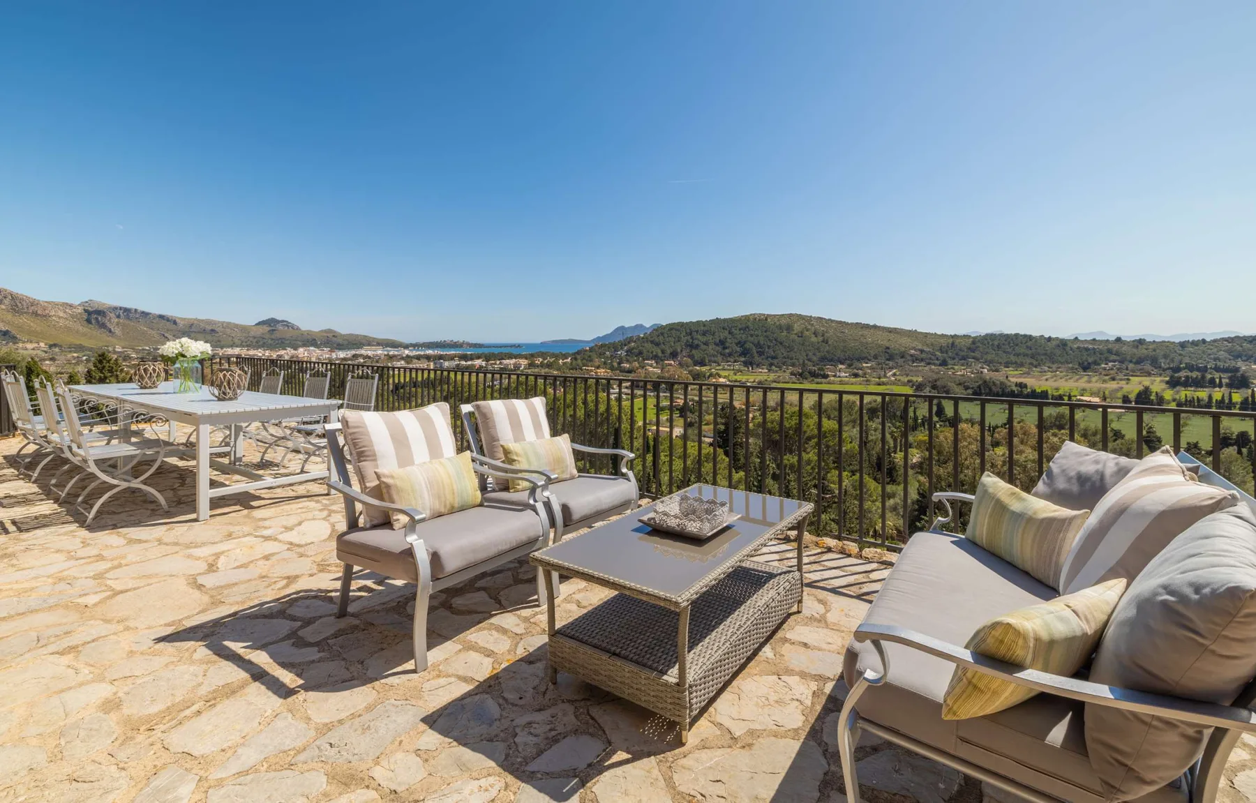 An outdoor terrace has cushioned seating and a dining table overlooking green hills.