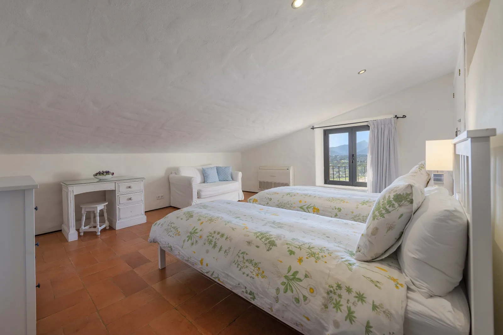 A bright bedroom with two beds, floral bedding, and a window.