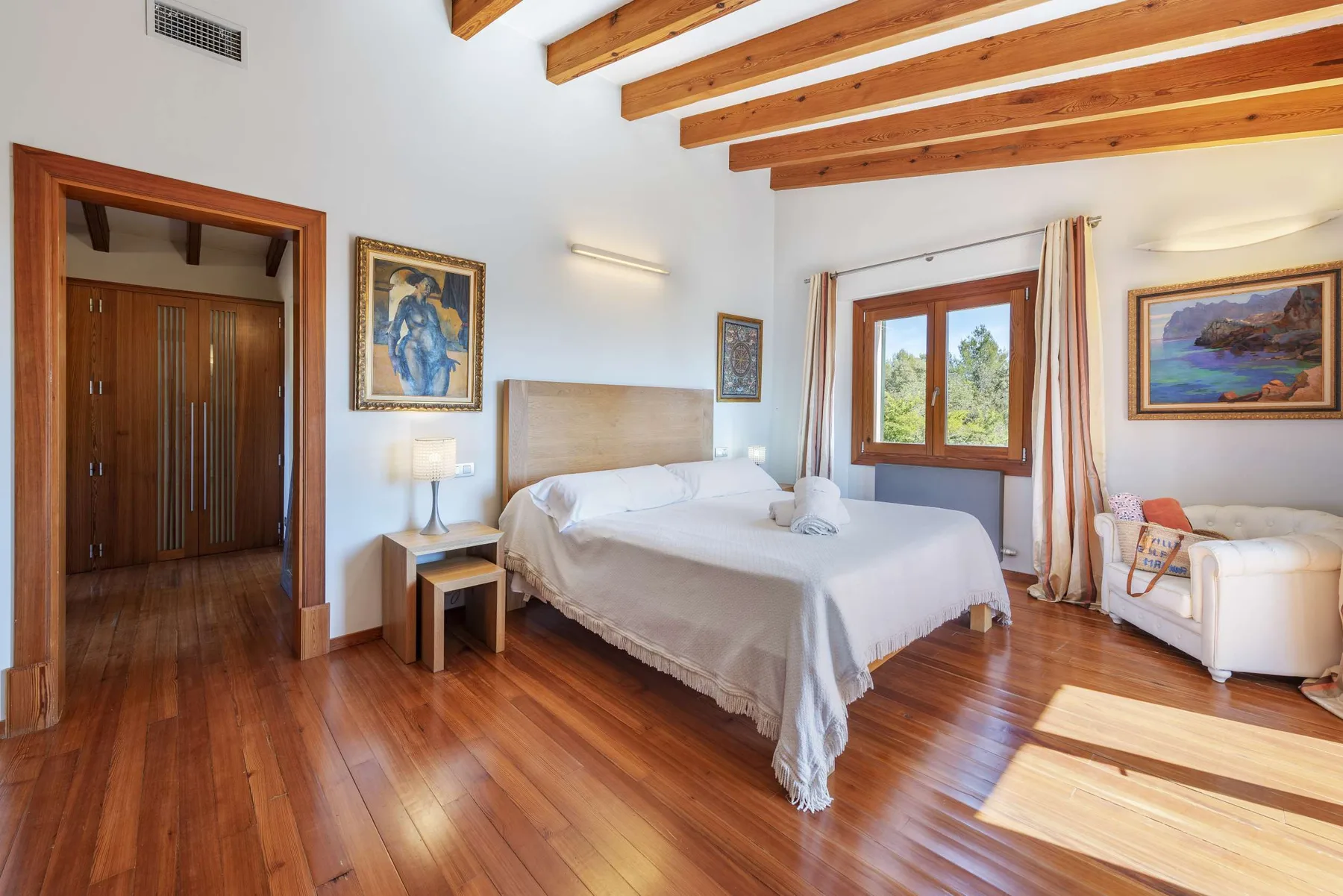 A bedroom with a large bed, wooden ceiling beams, and a sunlit window.