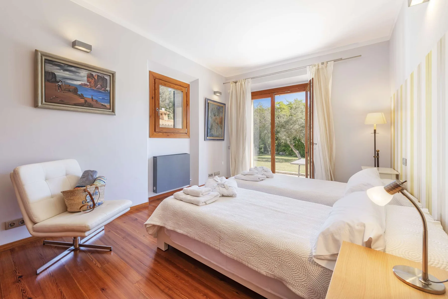 A bright bedroom with a large bed, a lounge chair, and glass doors opening onto greenery.