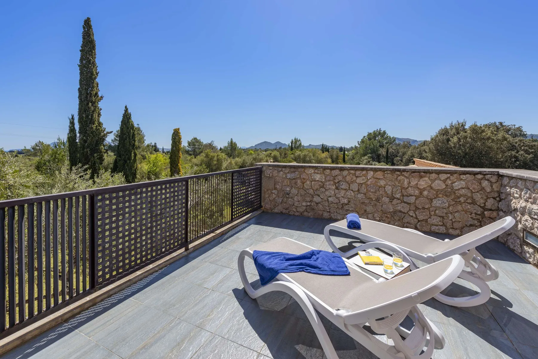 A sunny terrace has reclining loungers beside a stone wall, with trees and hills beyond.
