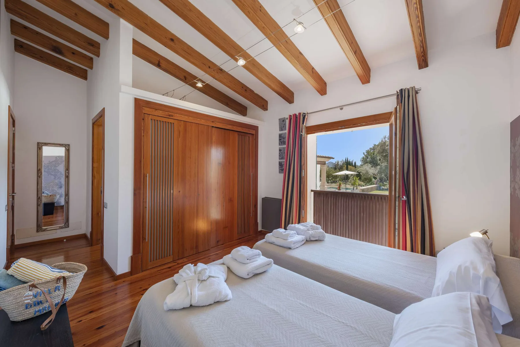 A bright bedroom with twin beds, exposed wooden ceiling beams, and an open balcony door.