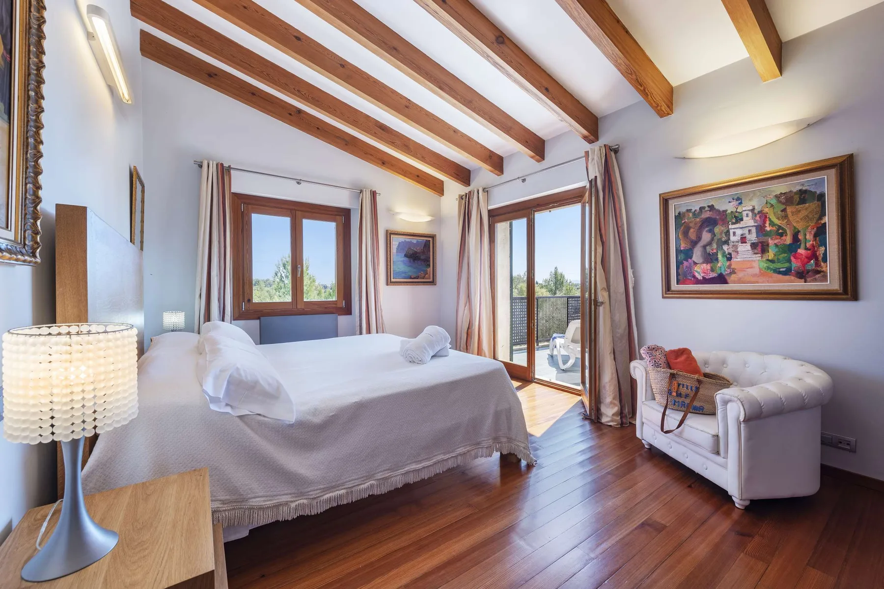A bright bedroom with a large bed, exposed wooden ceiling beams, and a small seating area.