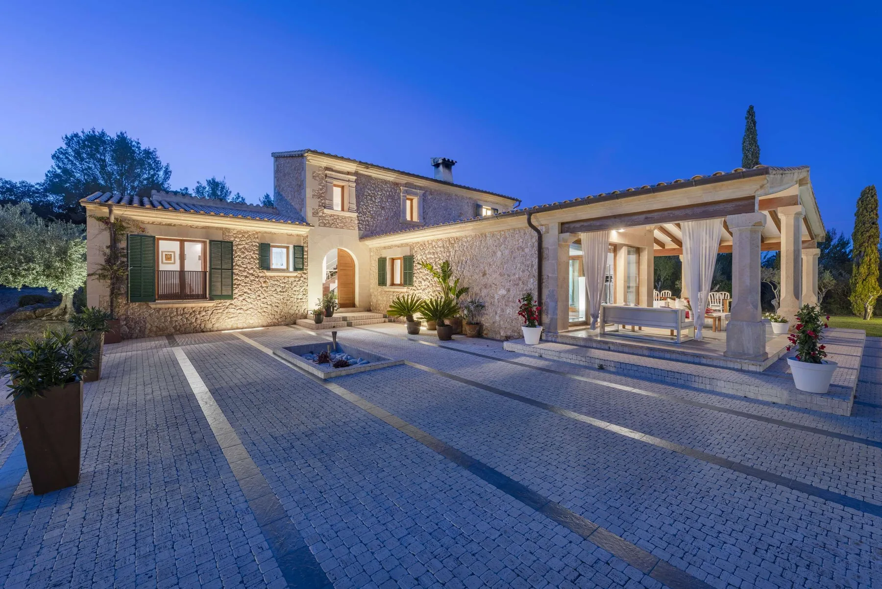 A stone house with a covered patio opens onto a paved courtyard at dusk.