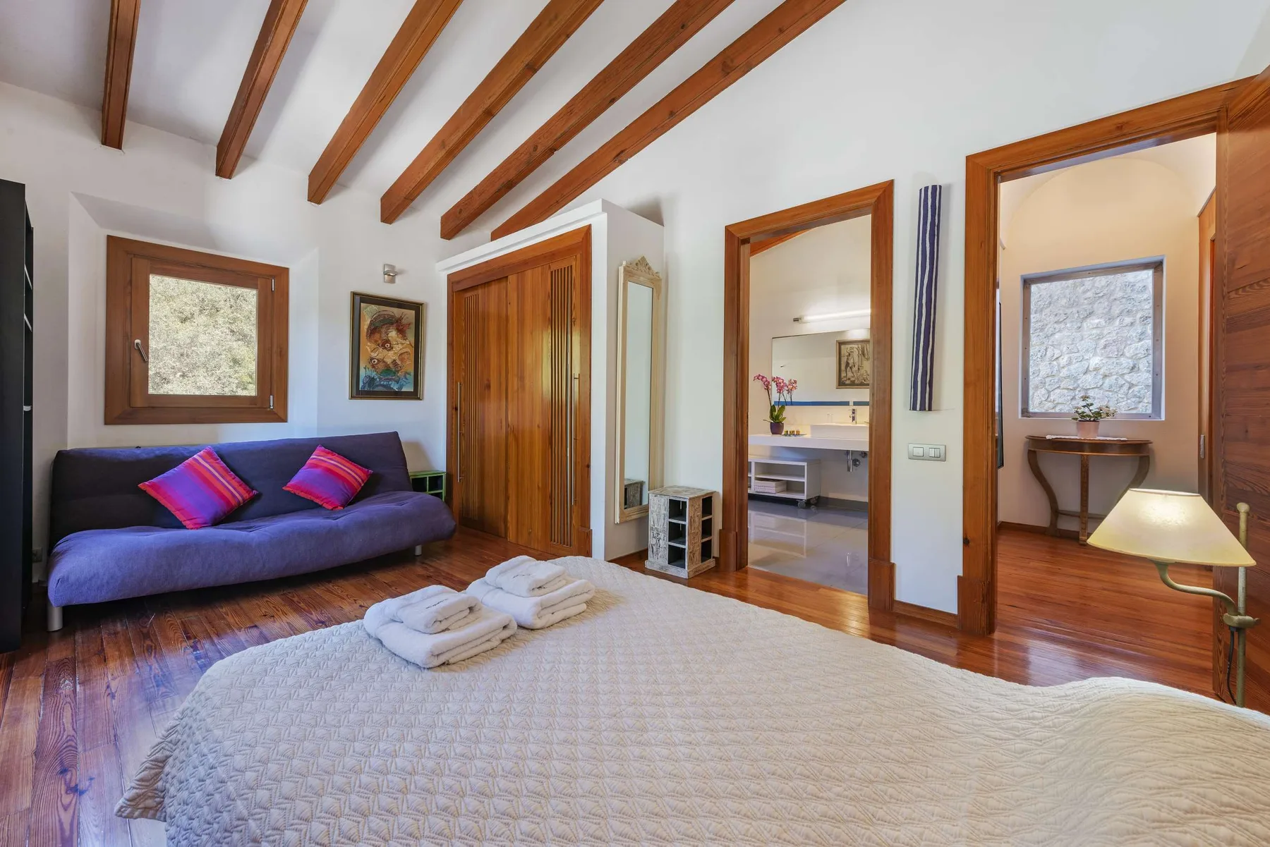 A bedroom with a bed, purple seating, and exposed wooden ceiling beams.