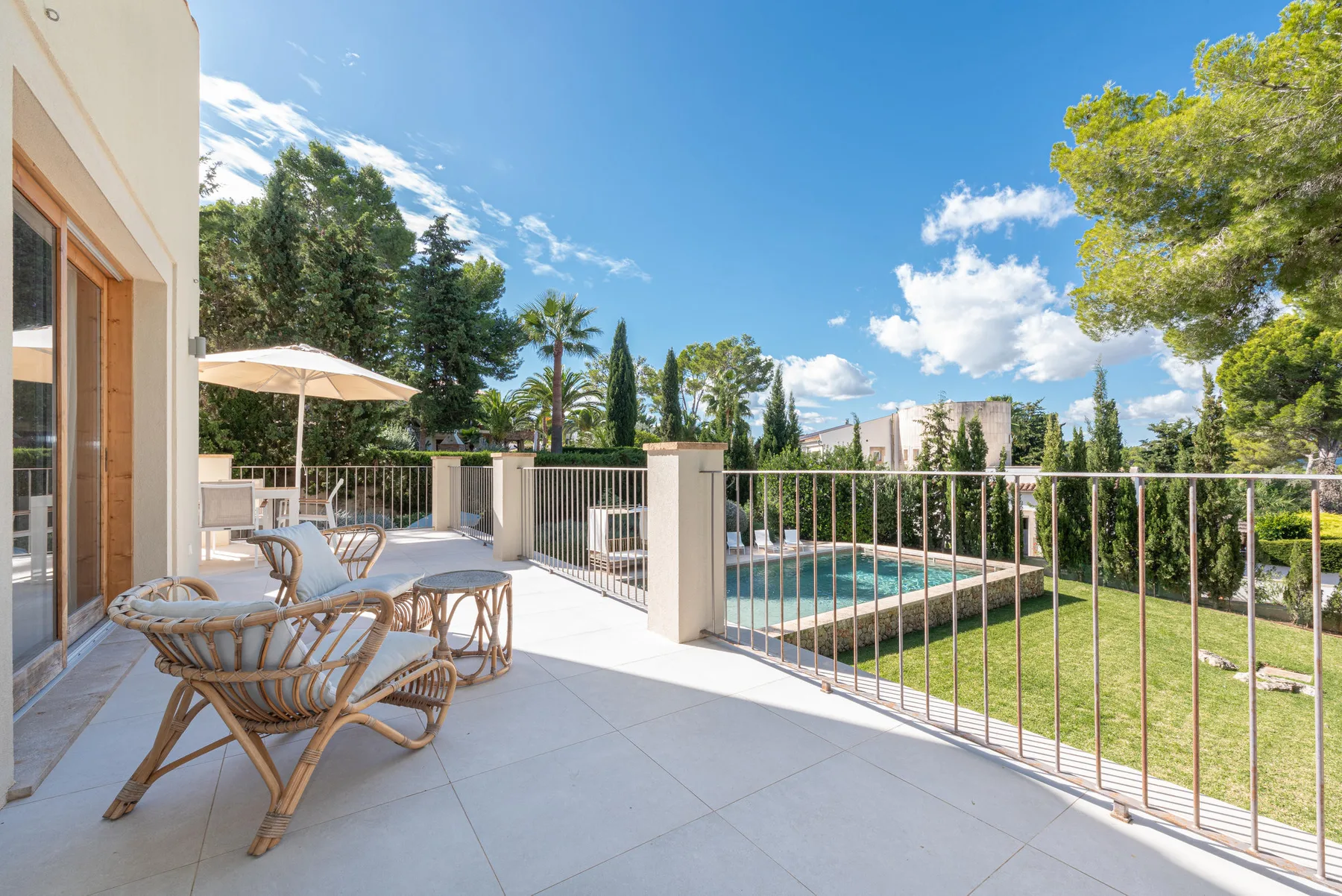A sunny patio with outdoor seating overlooks a swimming pool and garden.