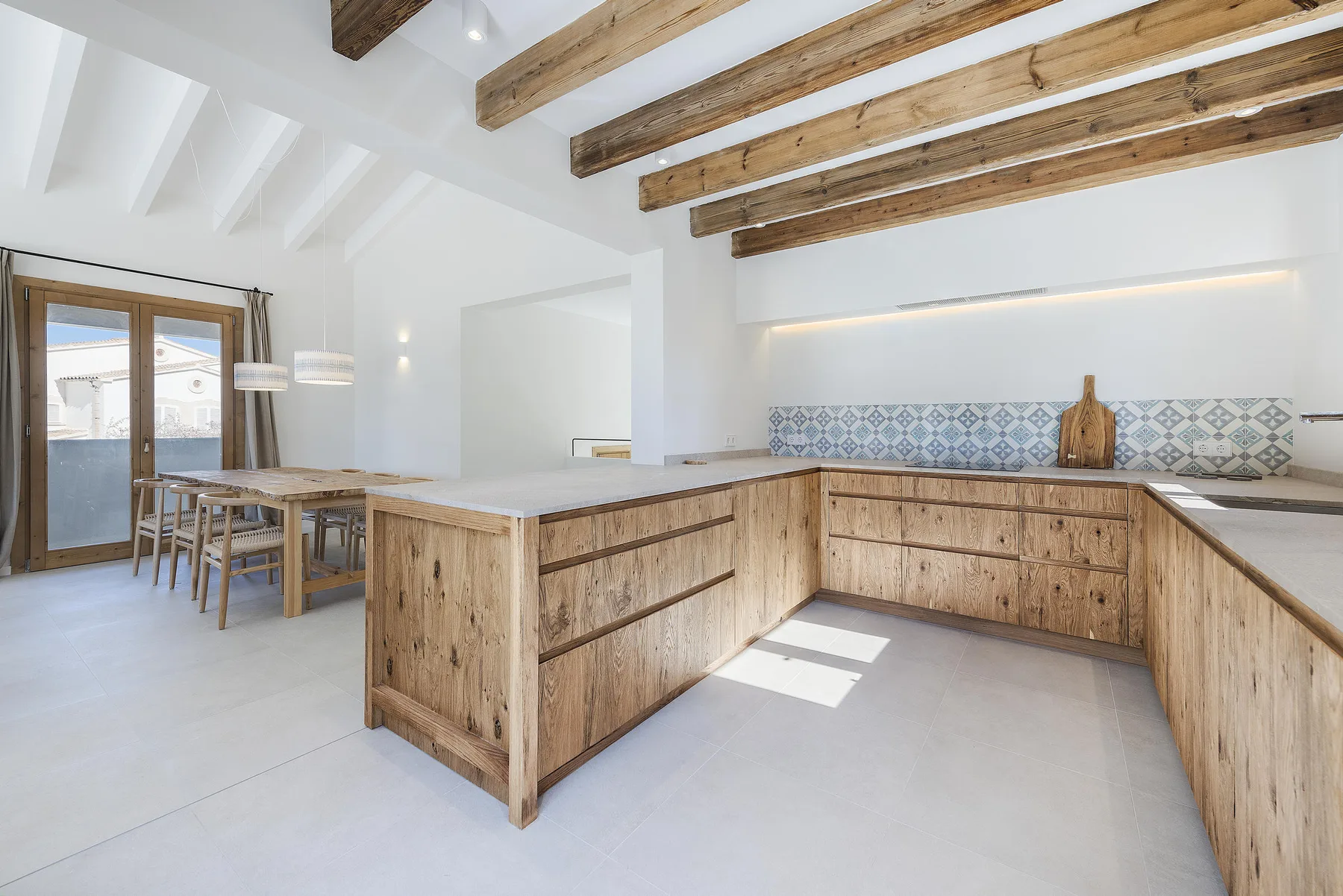 A bright kitchen with a large wooden island, dining table, and exposed ceiling beams.