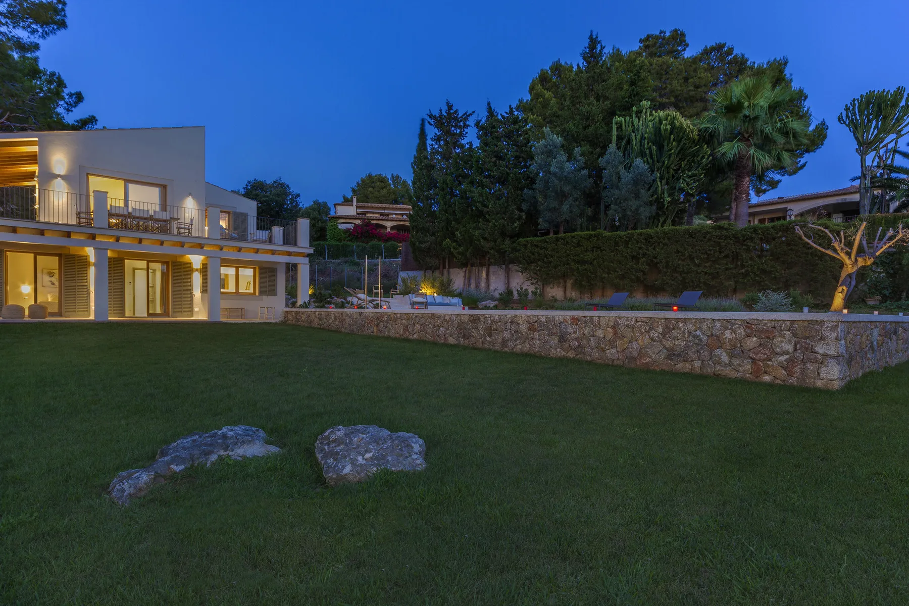 A modern house stands beside a broad lawn and stone retaining wall at dusk.