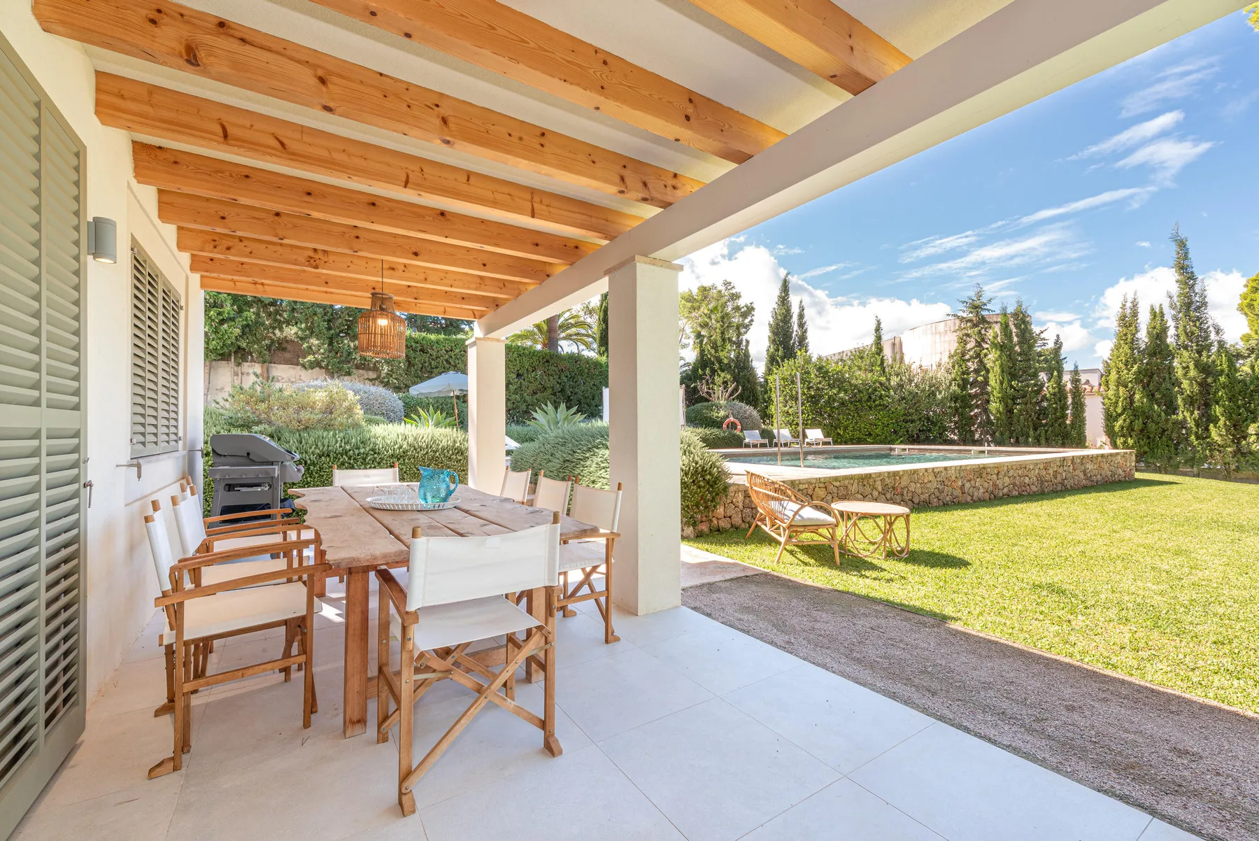 A covered patio has a dining table and seating beside a lawn and swimming pool.