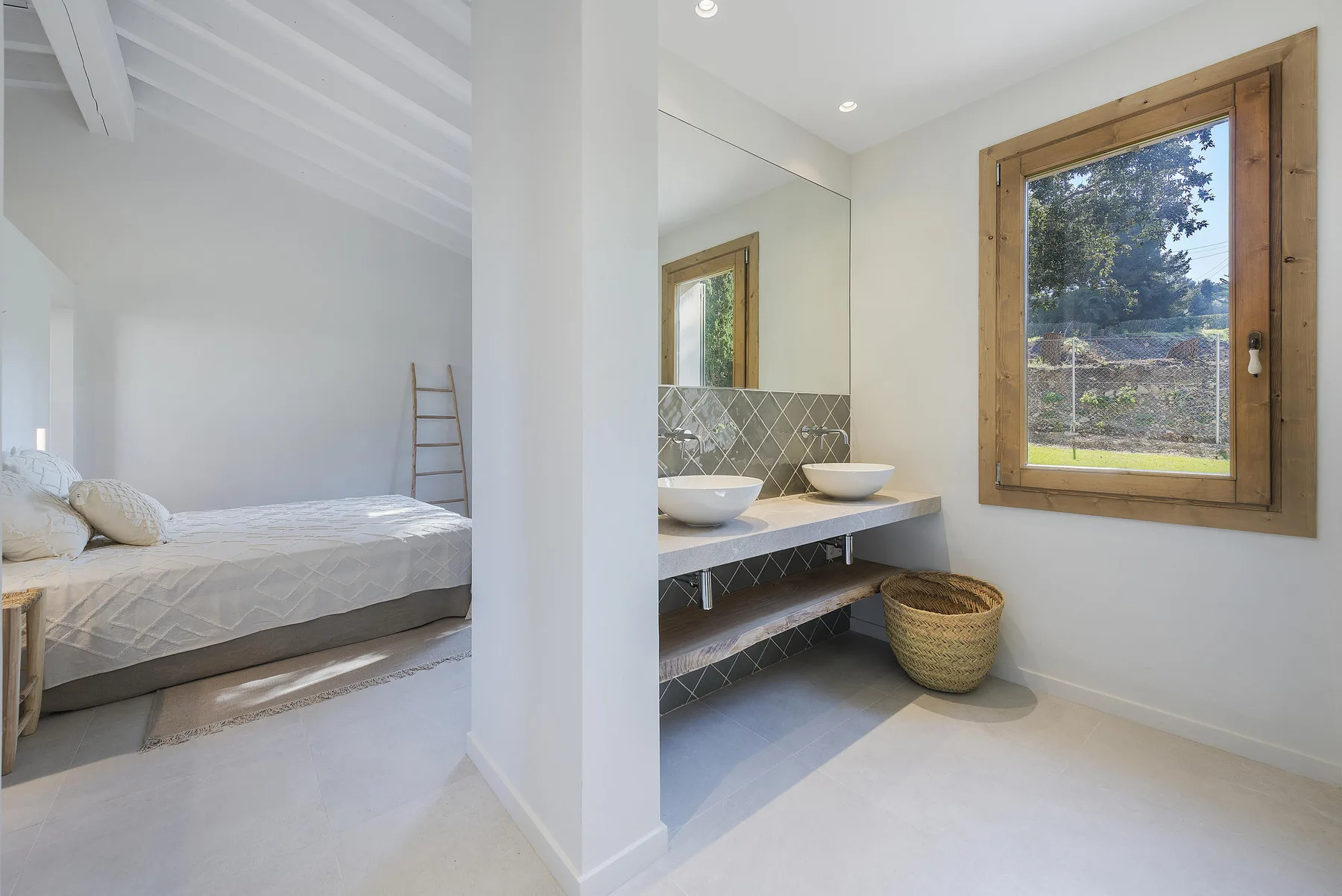 A bright bedroom opens onto a bathroom with a double vanity and a large window.
