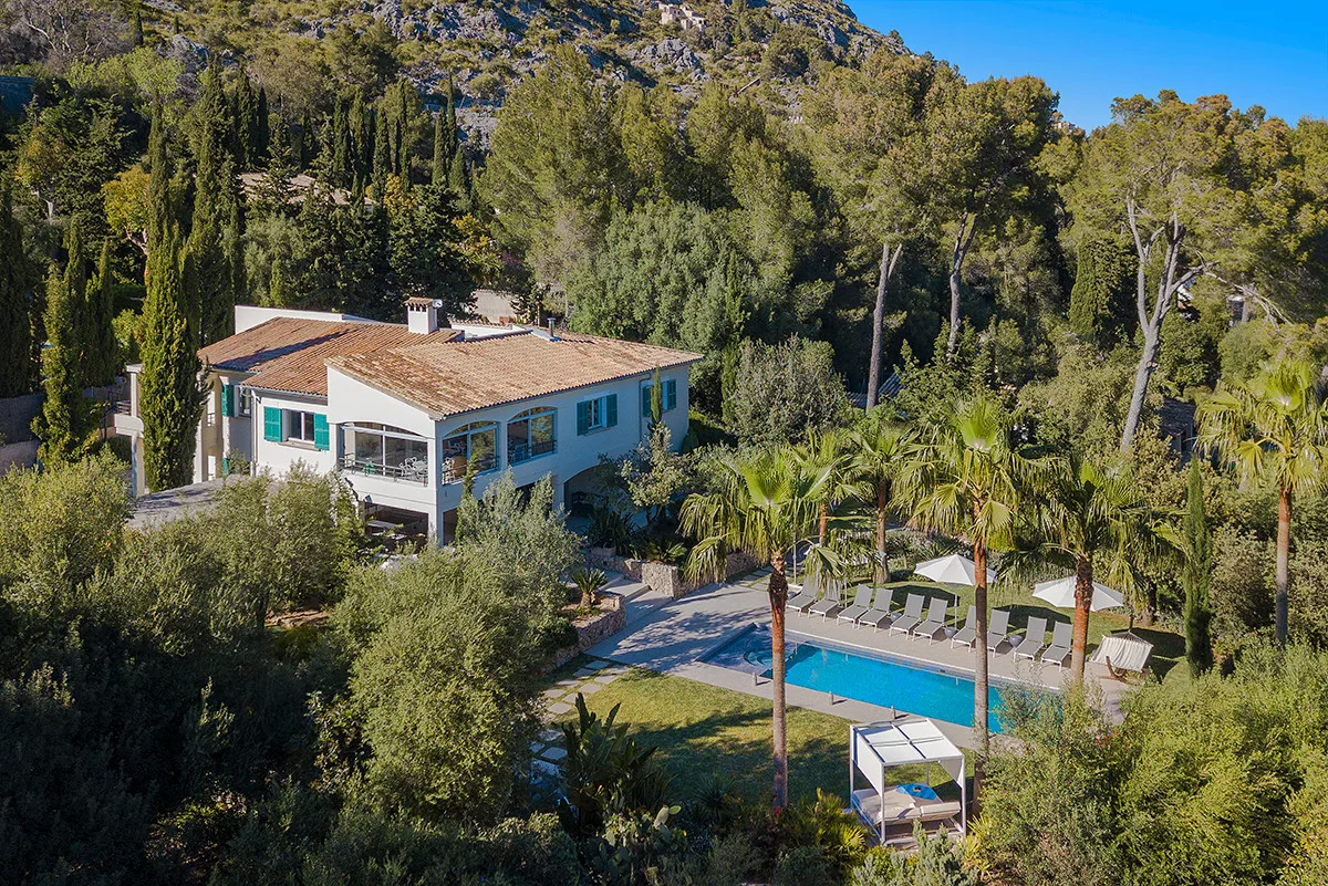 A house and swimming pool sit among dense trees on a wooded hillside.