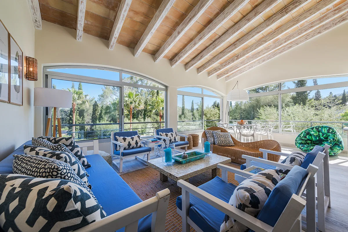 A bright sunroom with seating arranged around a low table and large windows.