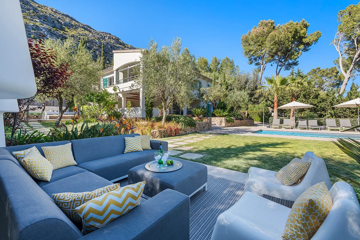 An outdoor lounge area with cushioned seating faces a garden and swimming pool.