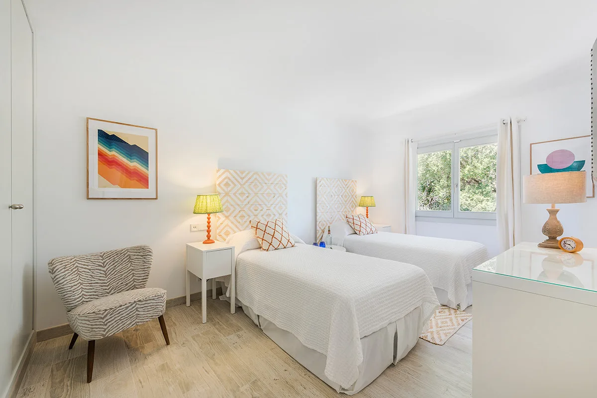 A bright bedroom with two beds, patterned chairs, and a window.