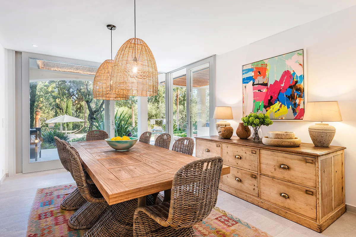 A bright dining area features a wooden table beside large windows overlooking greenery.
