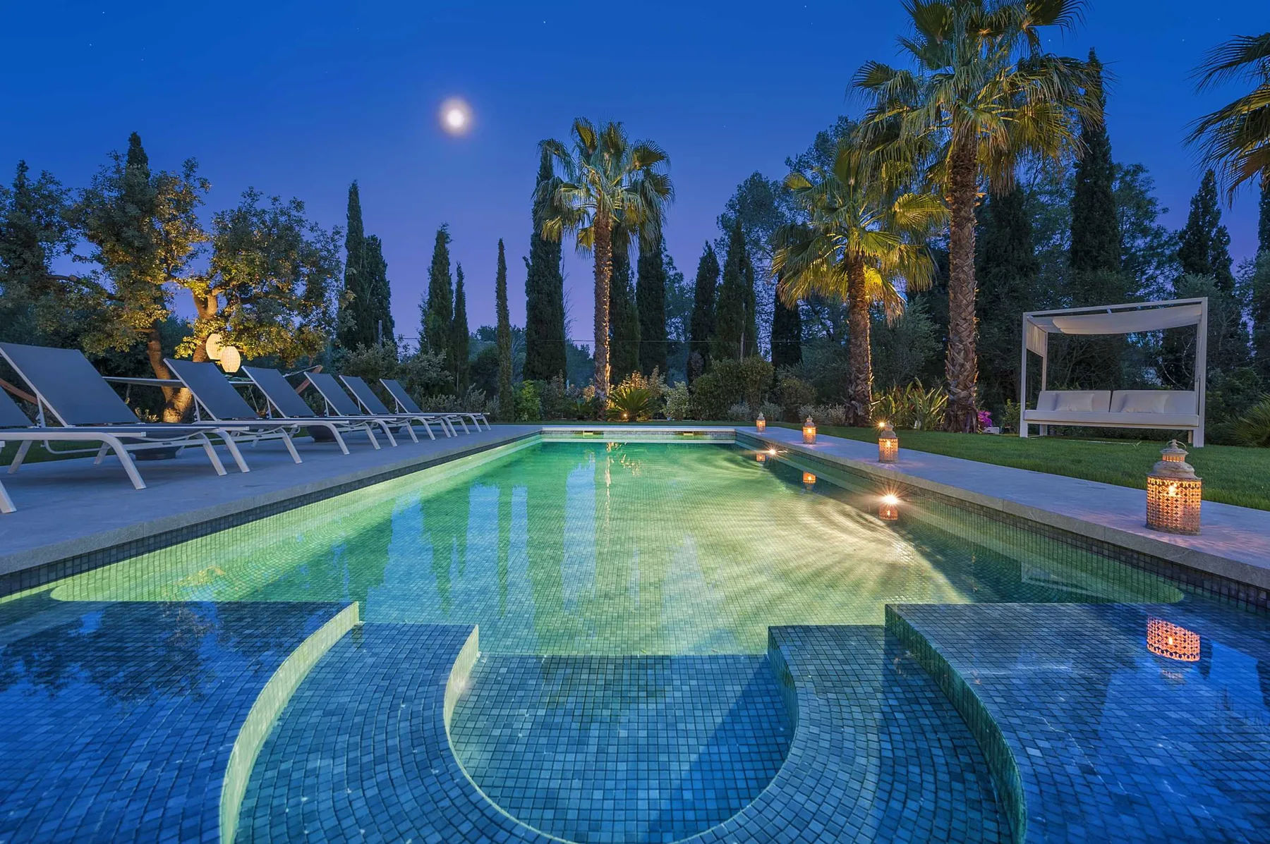 An outdoor swimming pool glows at dusk, with loungers and palm trees around it.