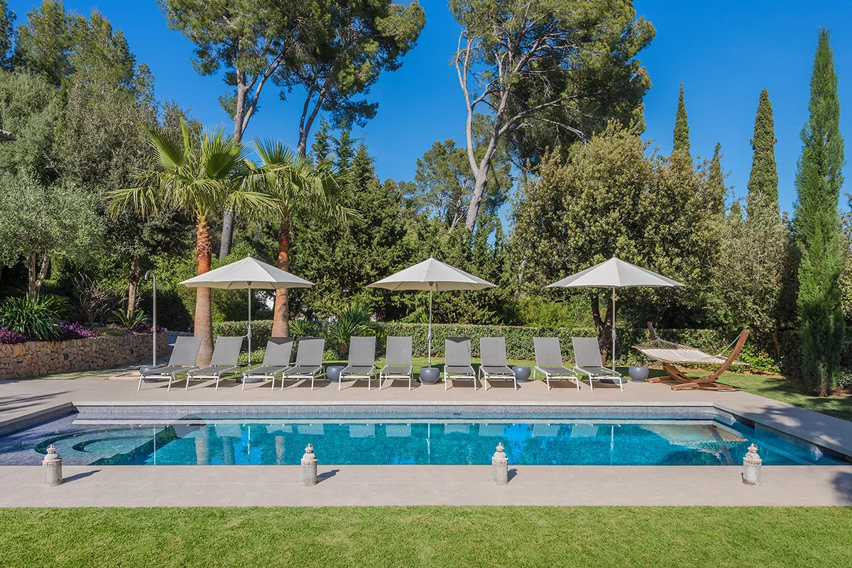 An outdoor swimming pool is bordered by lounge seating, umbrellas, and leafy trees.