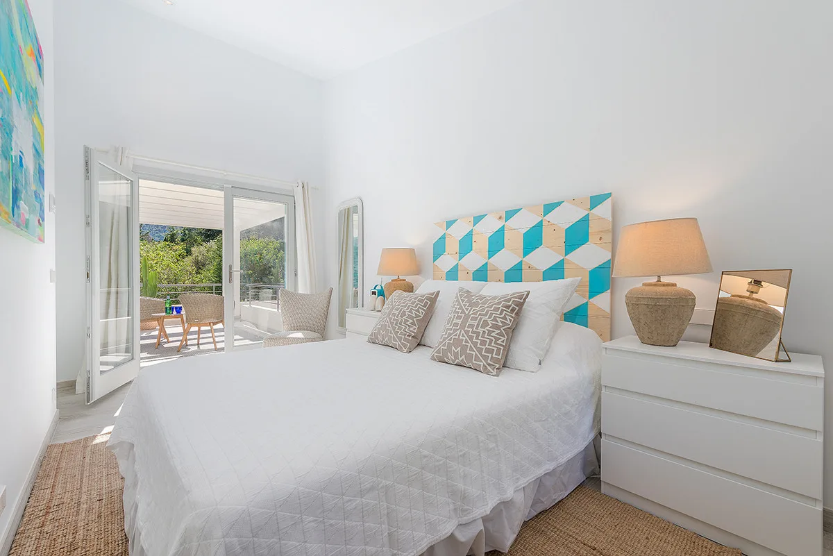 A bright bedroom with a white bed, patterned headboard, and glass doors opening to a balcony.