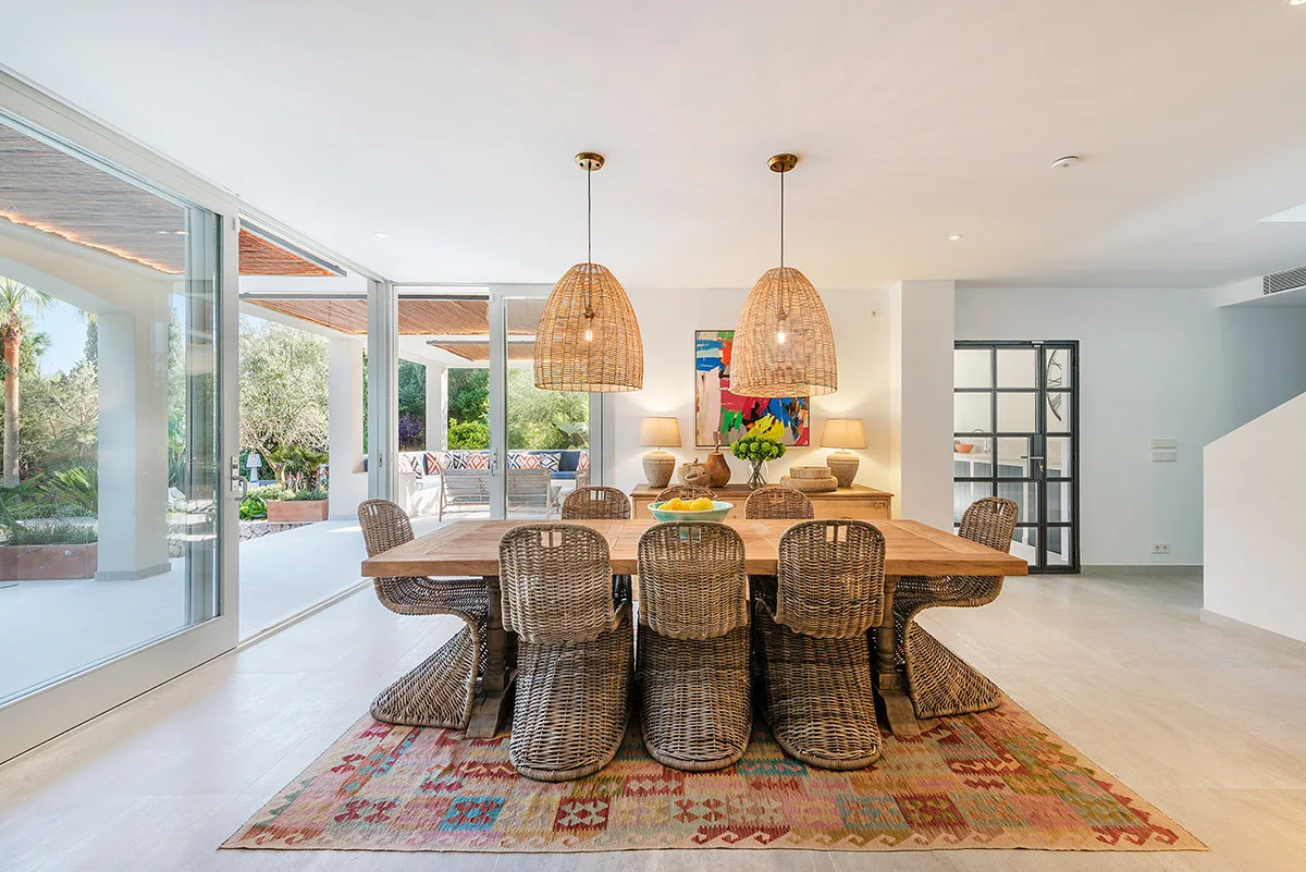 A dining area with a long table and woven seating beside large windows.