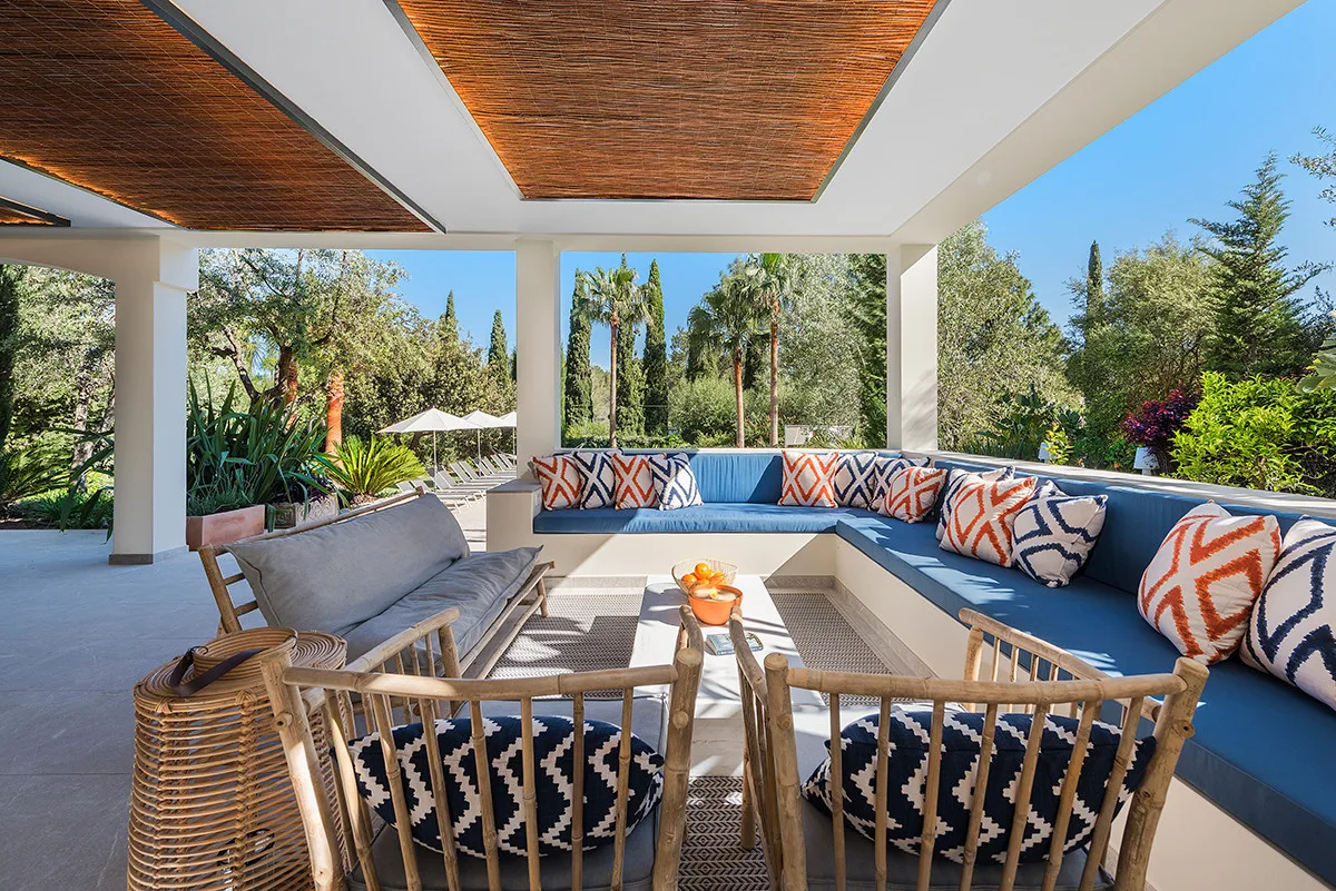 A covered outdoor seating area with cushioned benches and chairs overlooks a garden.