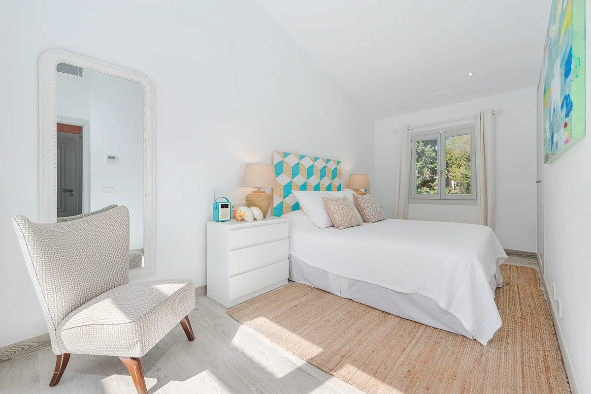 A bright bedroom with a neatly made bed, bedside dresser, and a window.