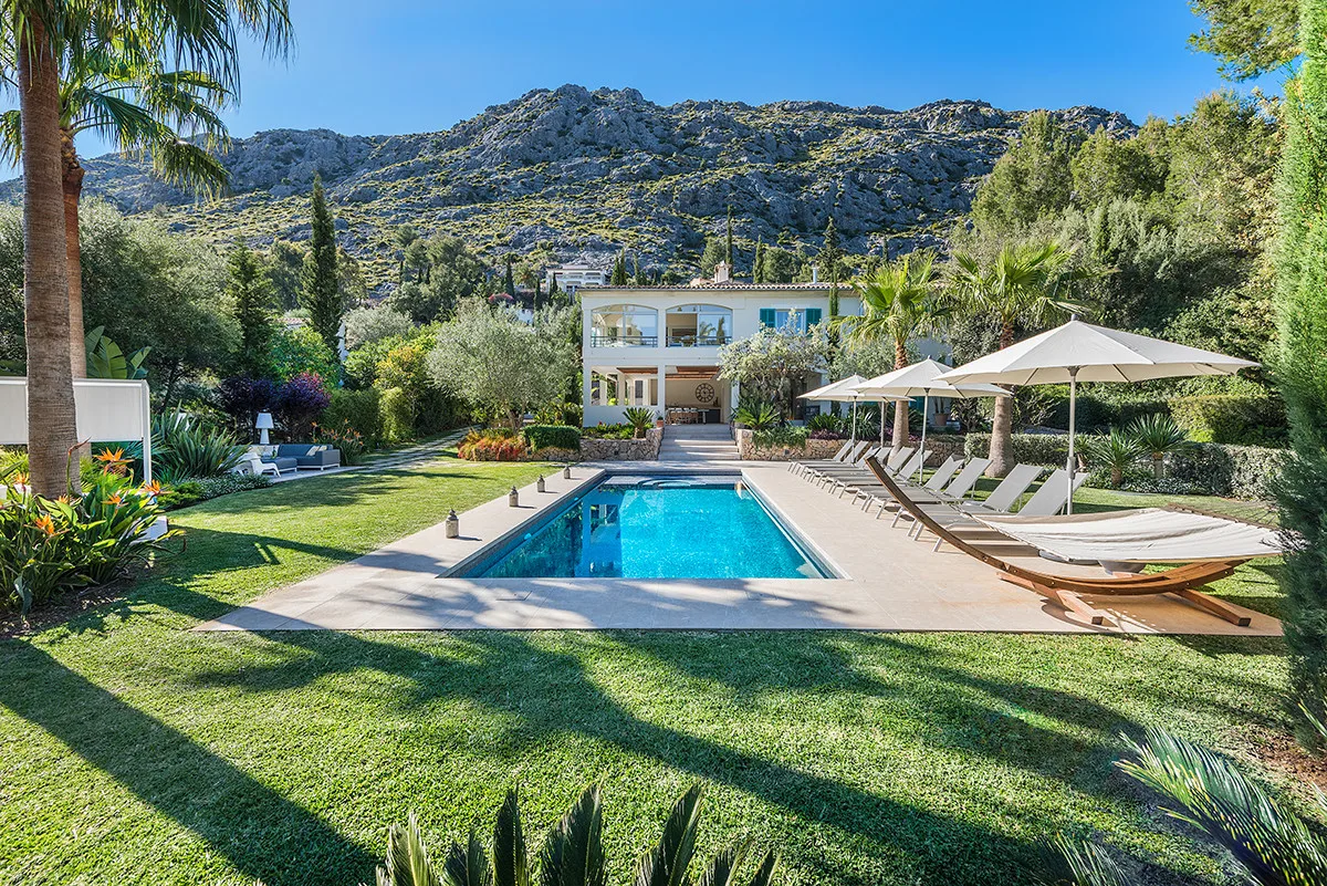 An outdoor swimming pool sits beside a landscaped lawn, with a building and mountains beyond.