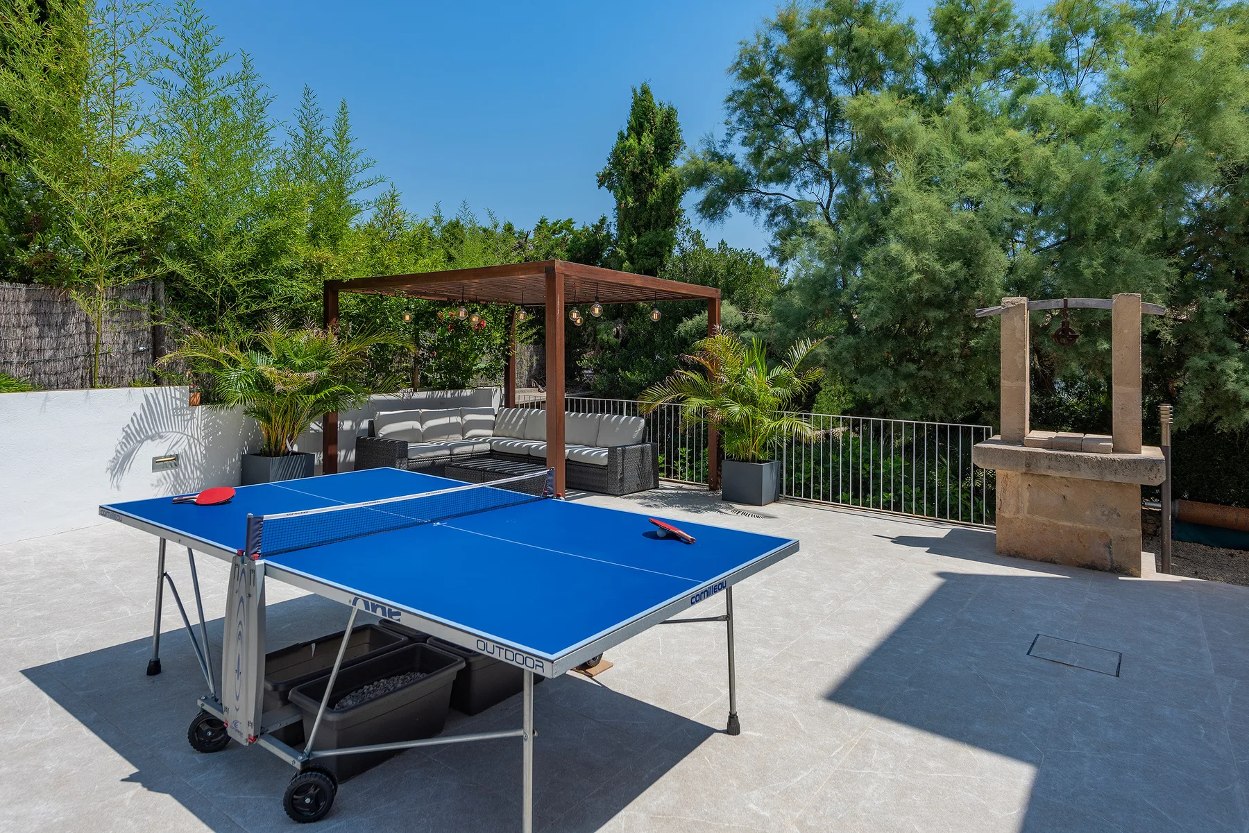 A blue ping-pong table stands on a sunny outdoor patio beside shaded seating.