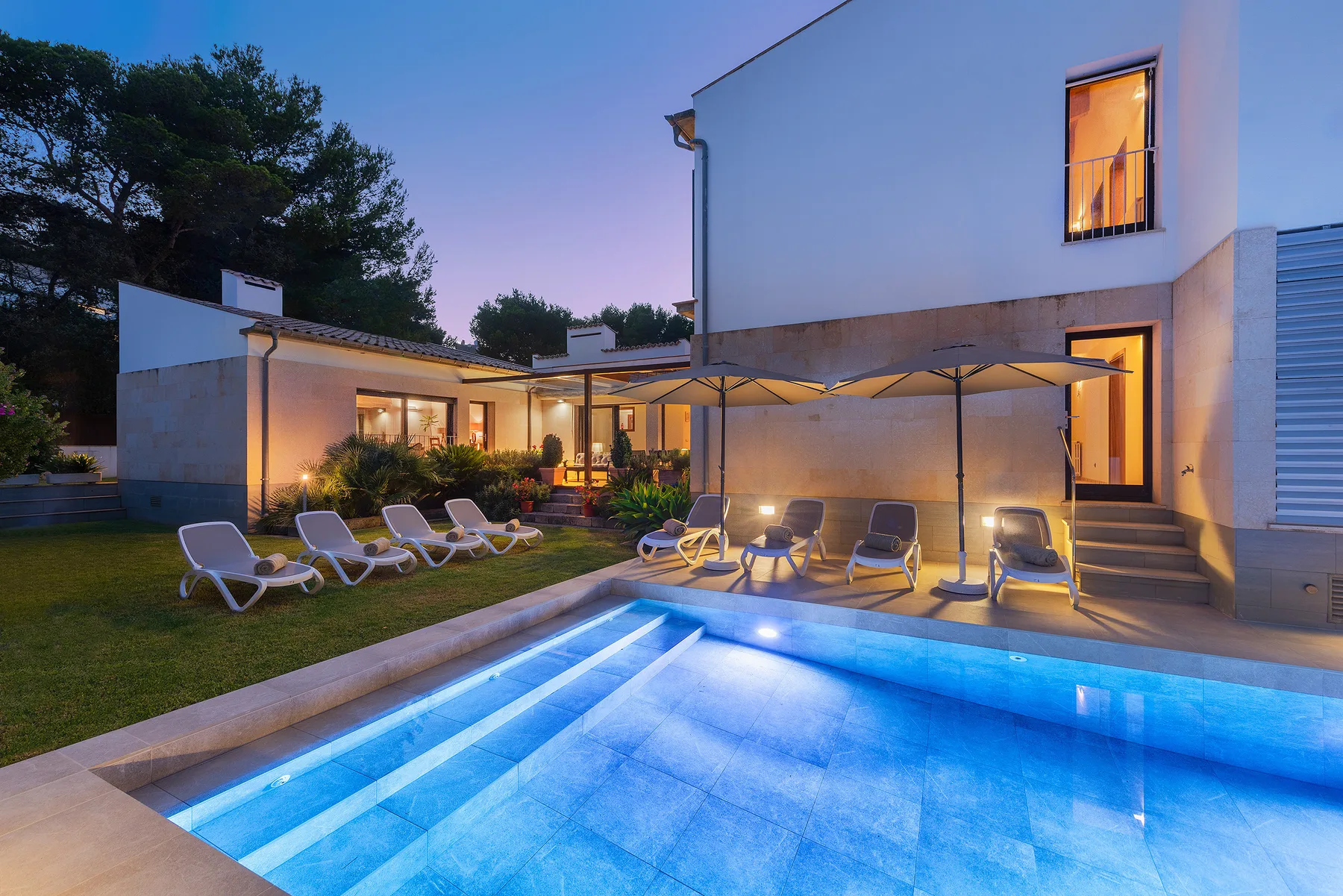 A modern house opens onto a lit swimming pool and a patio with outdoor seating.