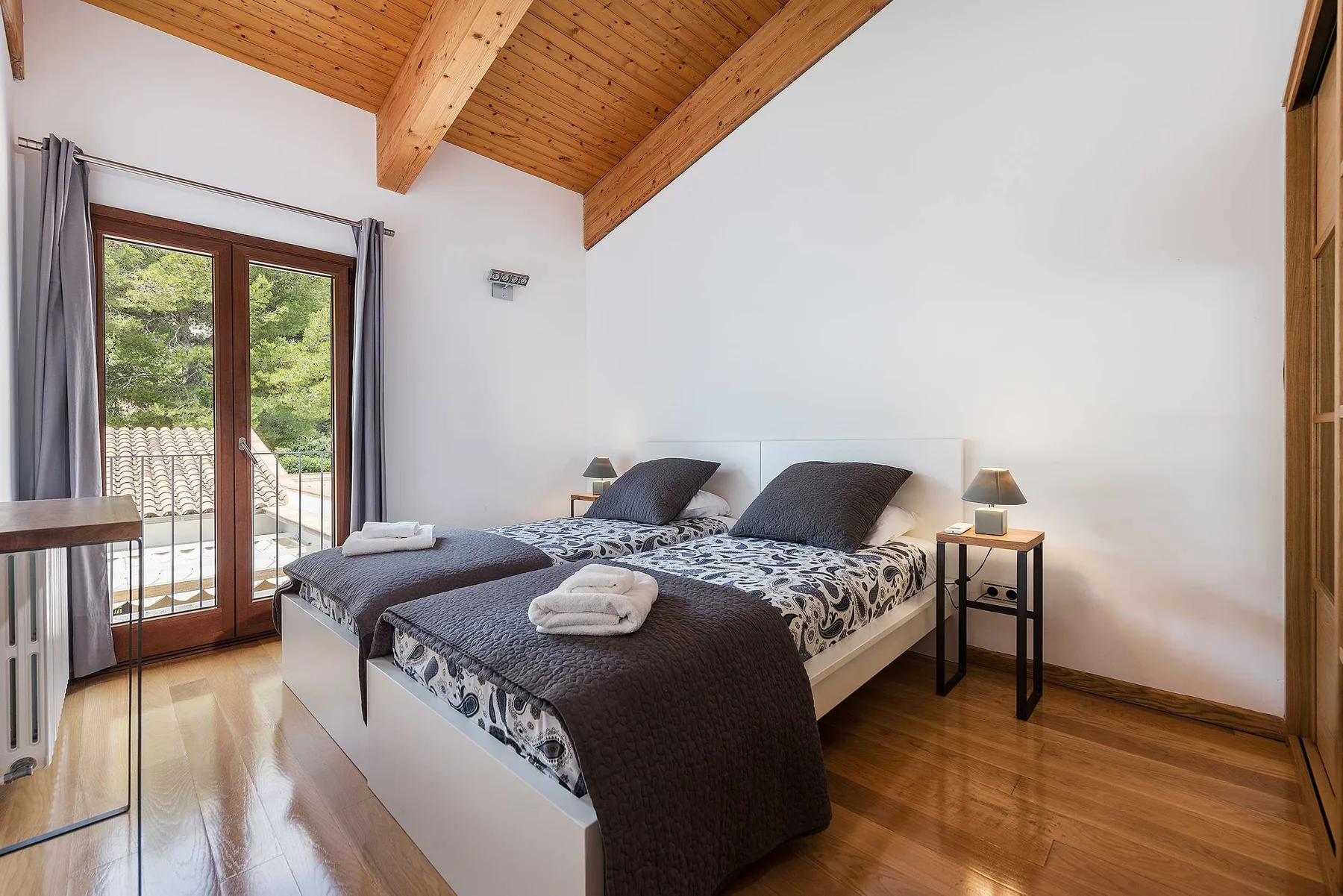 A bright bedroom with two beds beside tall glass doors opening onto a balcony.