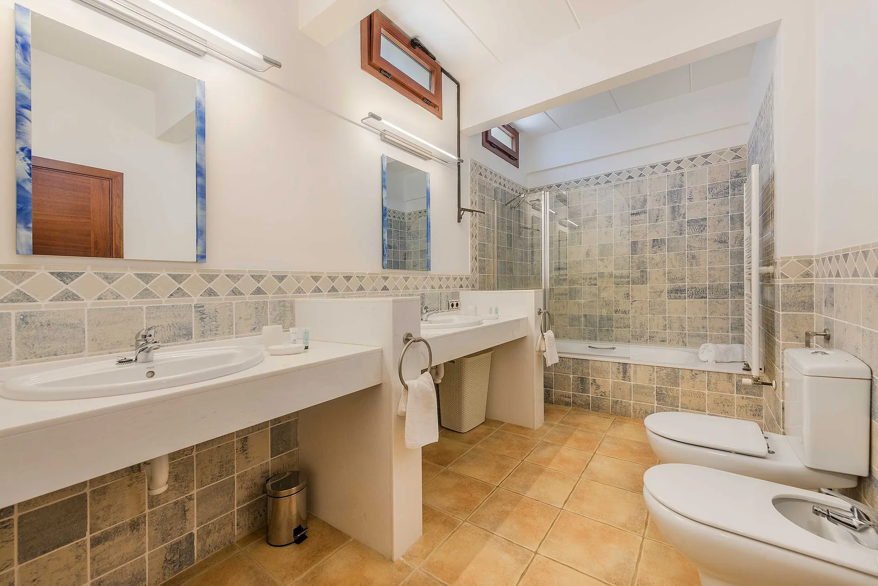 A tiled bathroom with a long sink counter, mirror, bathtub, and toilet.