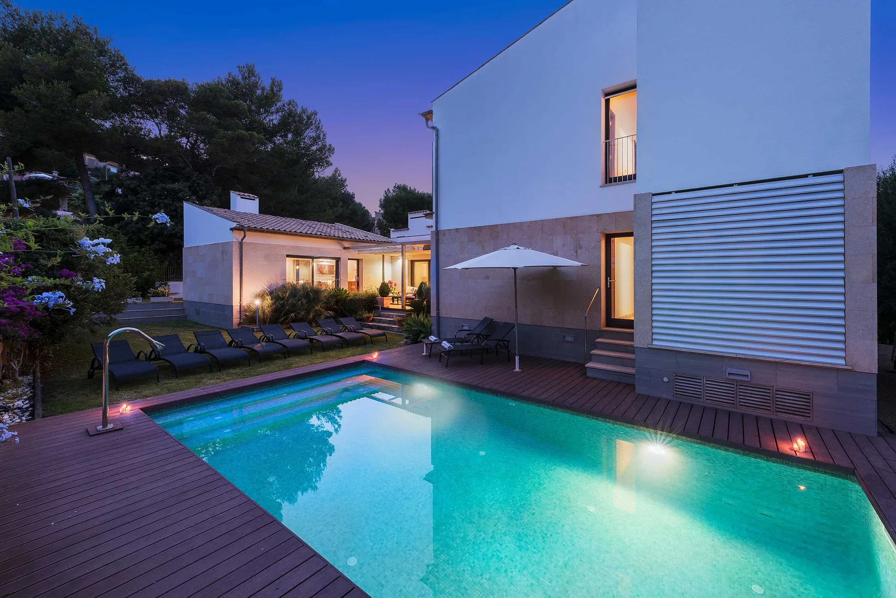 An outdoor swimming pool beside a modern house, with a wooden deck and lounge seating.