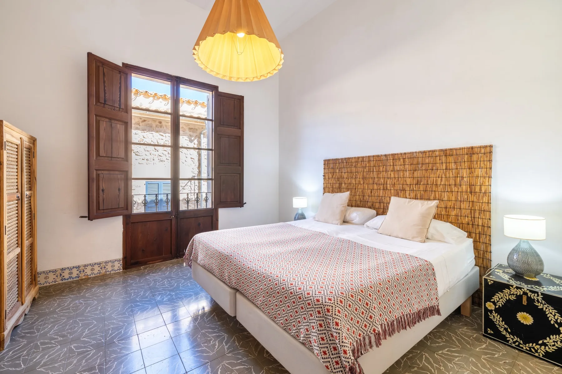 Un dormitorio con una colcha estampada y una ventana alta con contraventanas de madera.