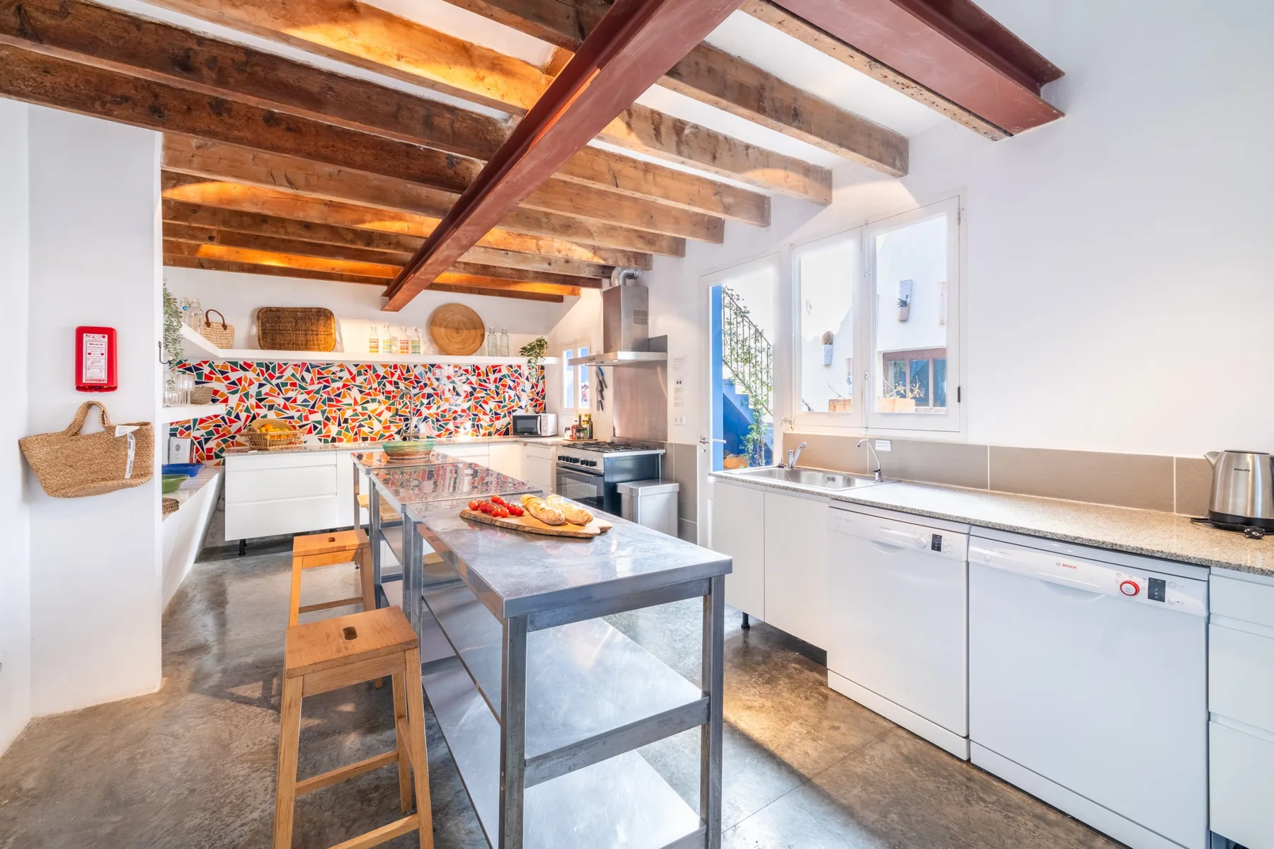 Una cocina luminosa con una mesa de trabajo central, armarios blancos y vigas de madera expuestas.
