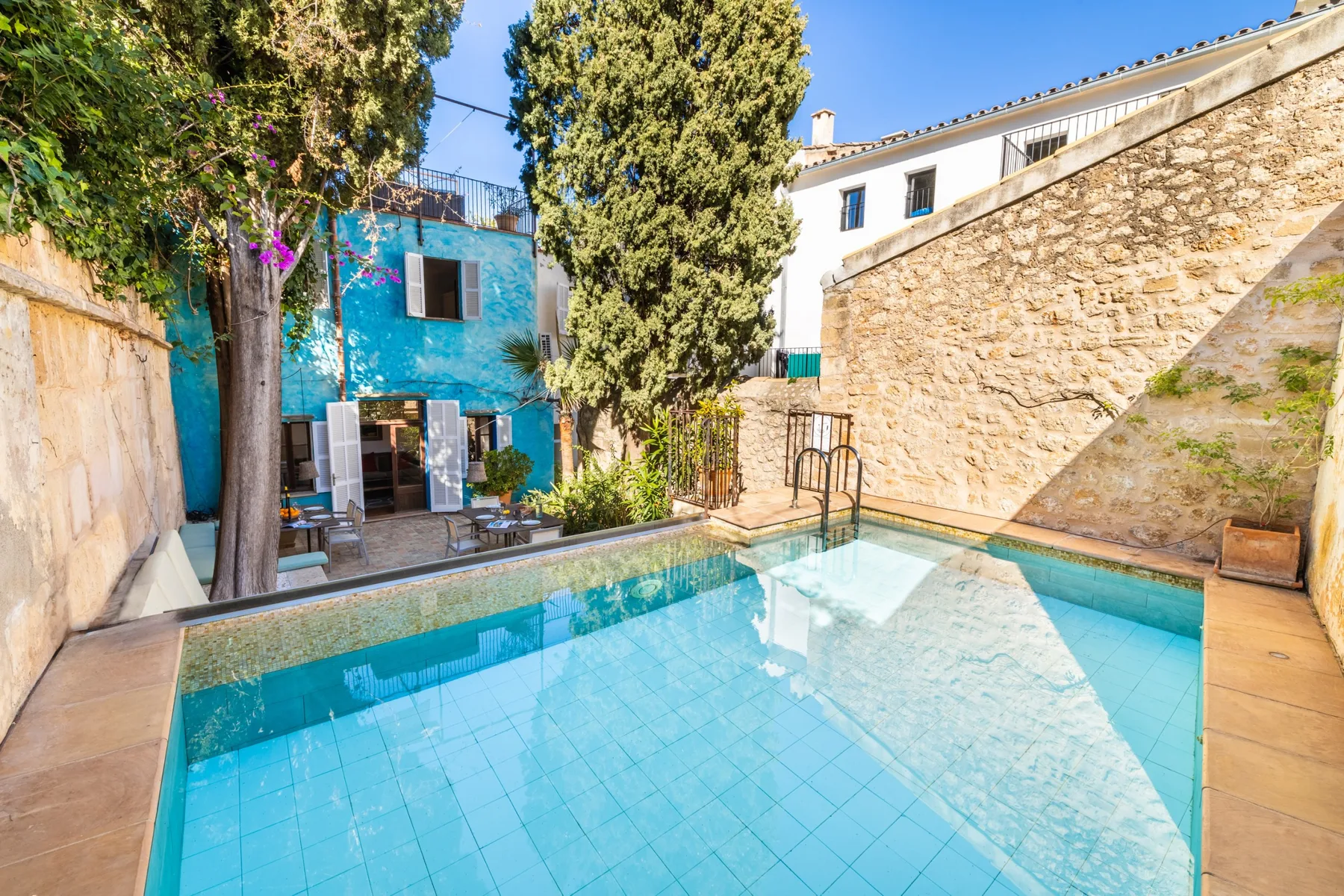 Una piscina exterior de azulejos está junto a un edificio azul y muros de piedra.