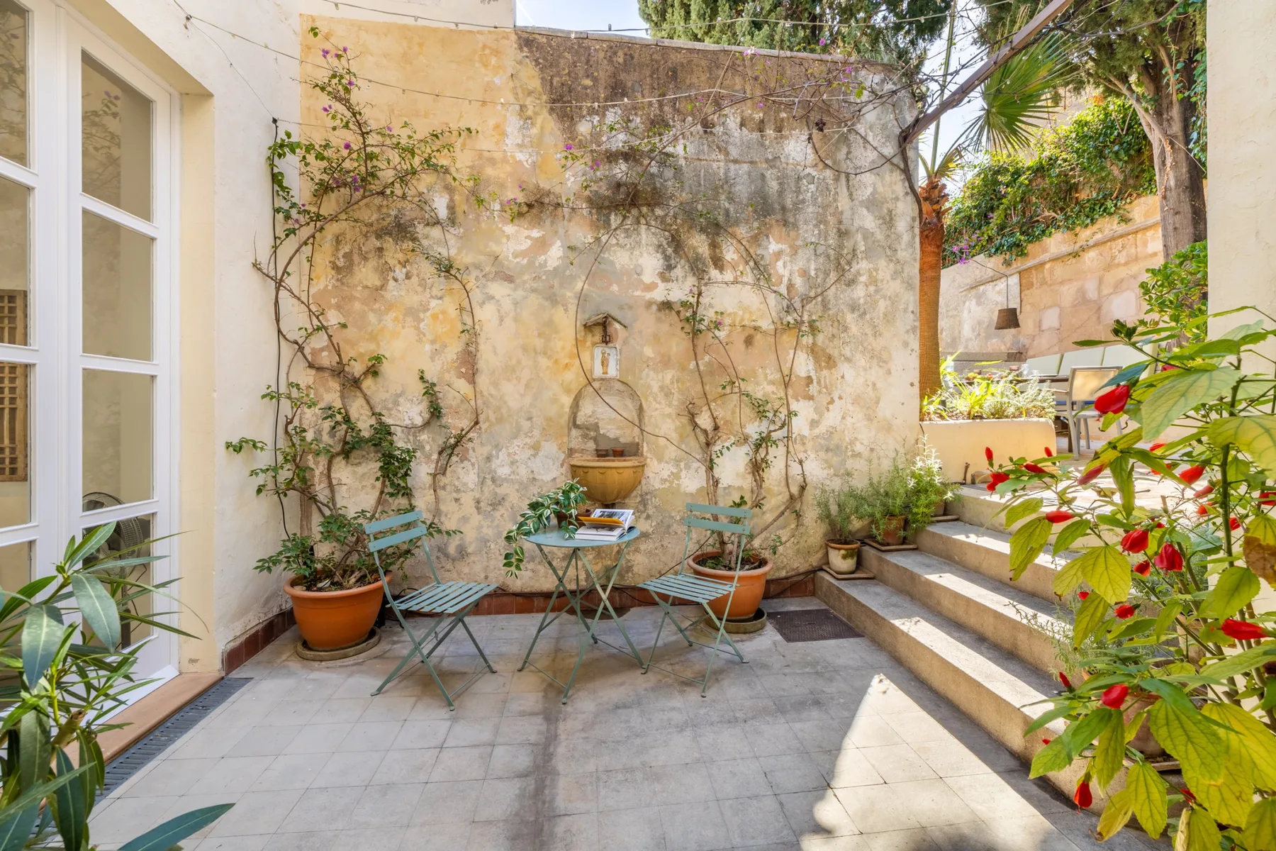 Un patio de piedra soleado con una pequeña zona de estar al aire libre y plantas en macetas.