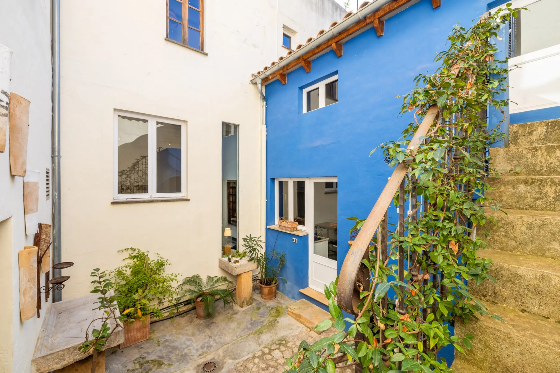 Un pequeño patio con una pared exterior azul, plantas en macetas y escalones de piedra.