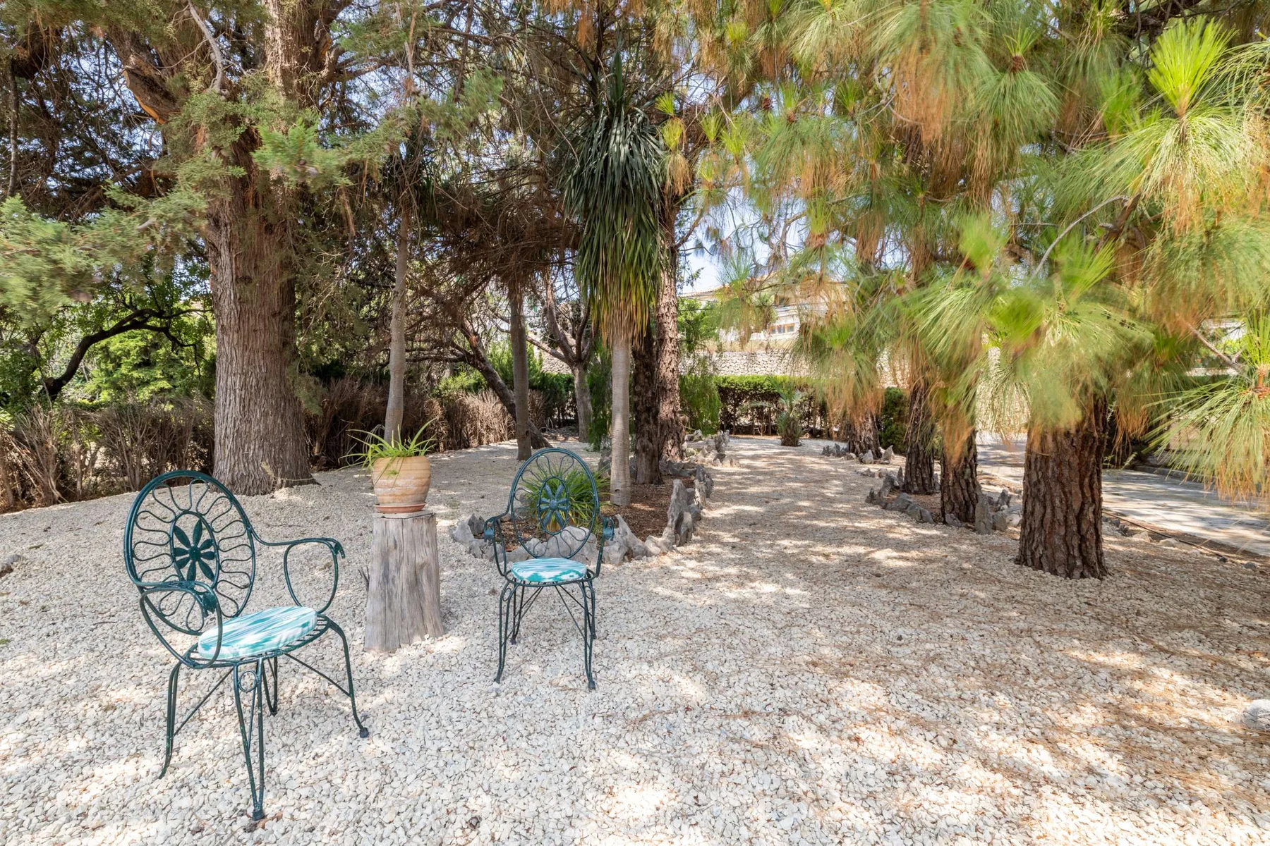 A shaded outdoor garden with gravel ground, trees, and metal seating.