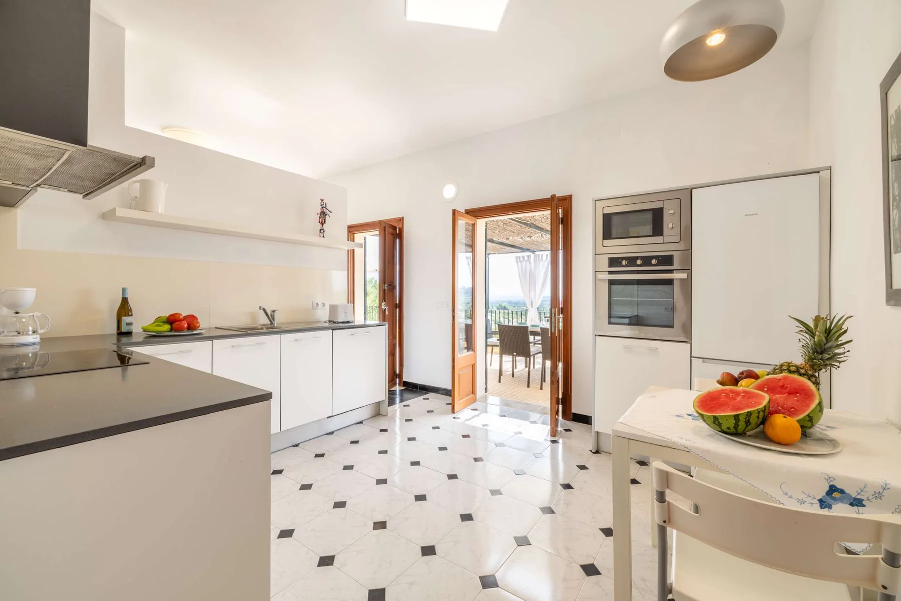 A bright kitchen with white cabinetry, a tiled floor, and a dining area beyond.