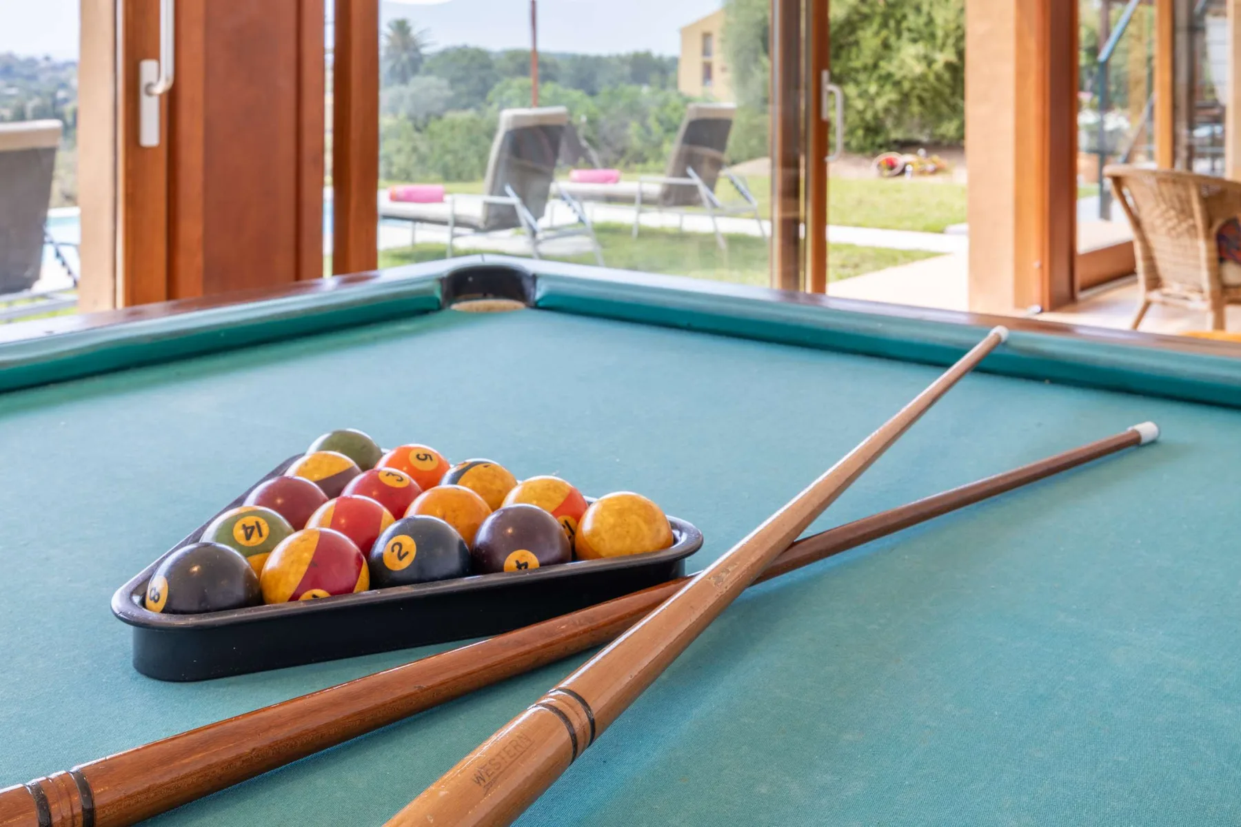A pool table with a rack of balls and two cues sits beside large windows.