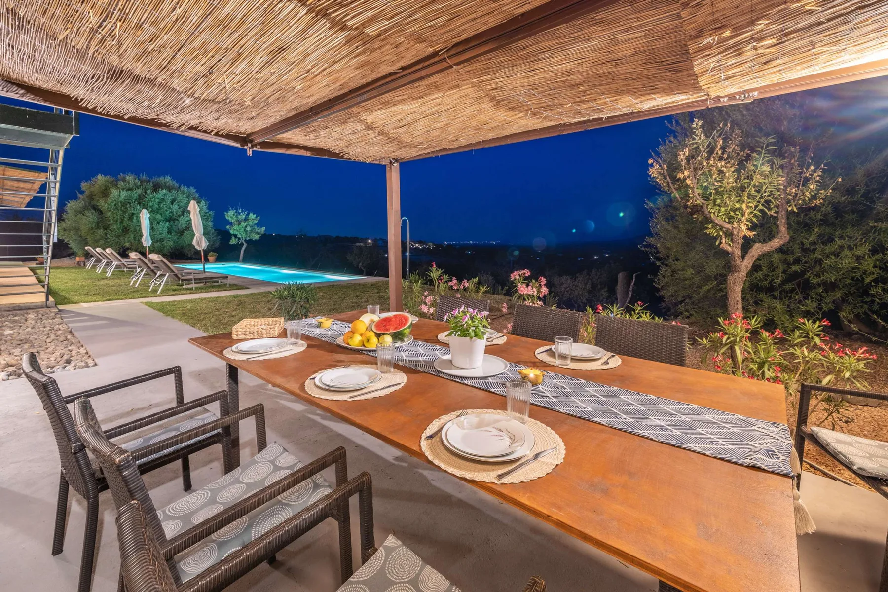 A covered outdoor dining area overlooks a swimming pool and garden at night.
