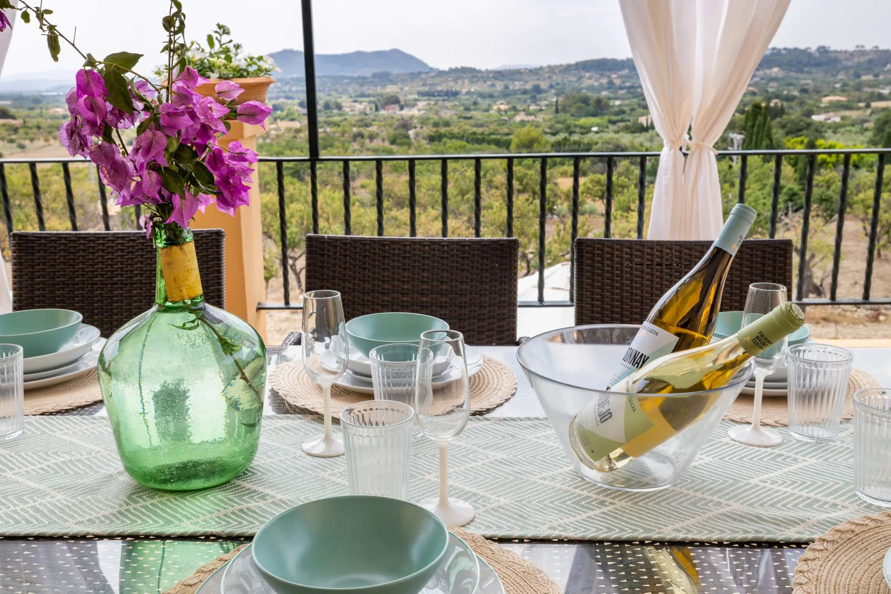 A dining table set with glassware and flowers overlooks a green valley.