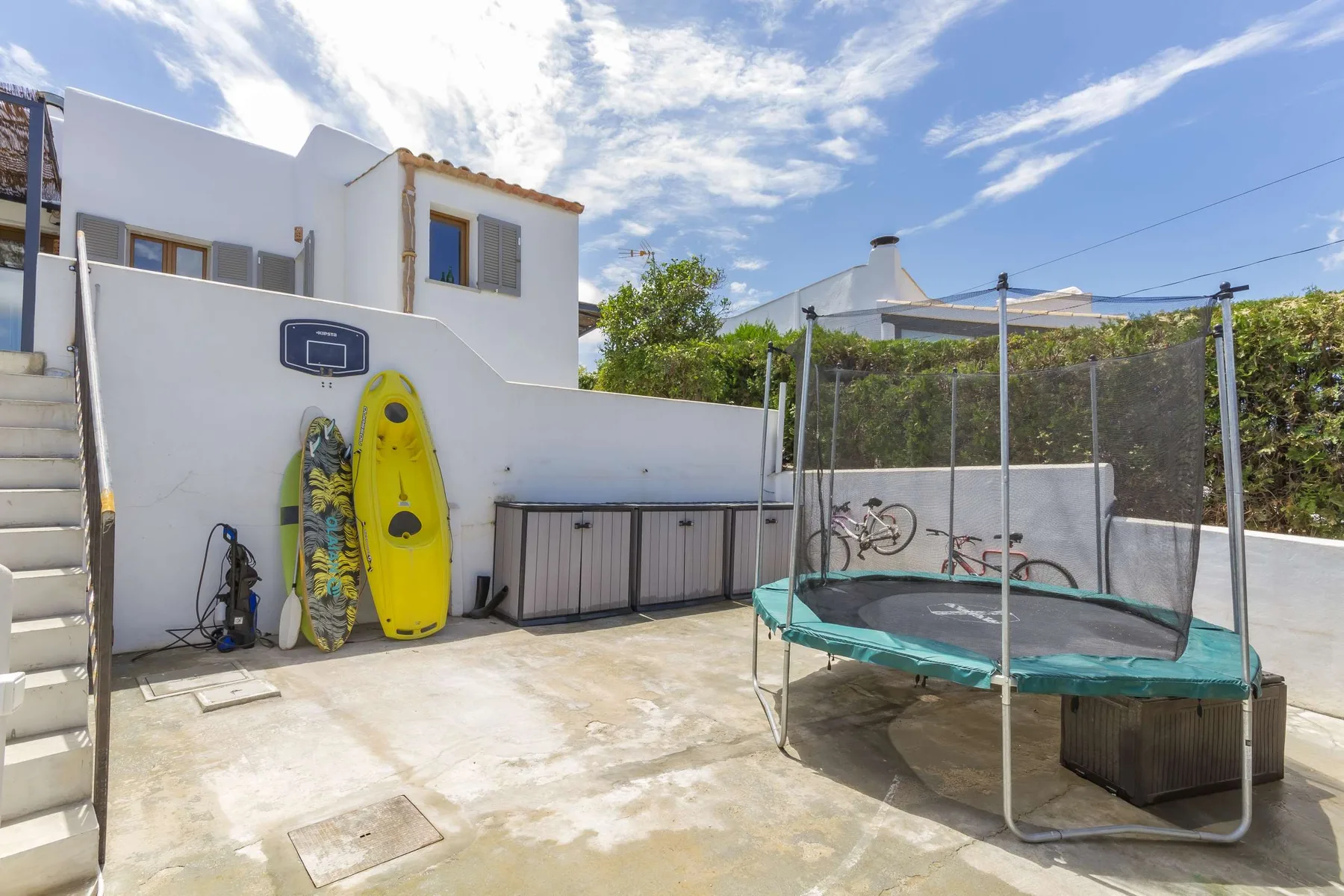 A sunny outdoor recreation area with a trampoline, surfboards, and a basketball hoop.