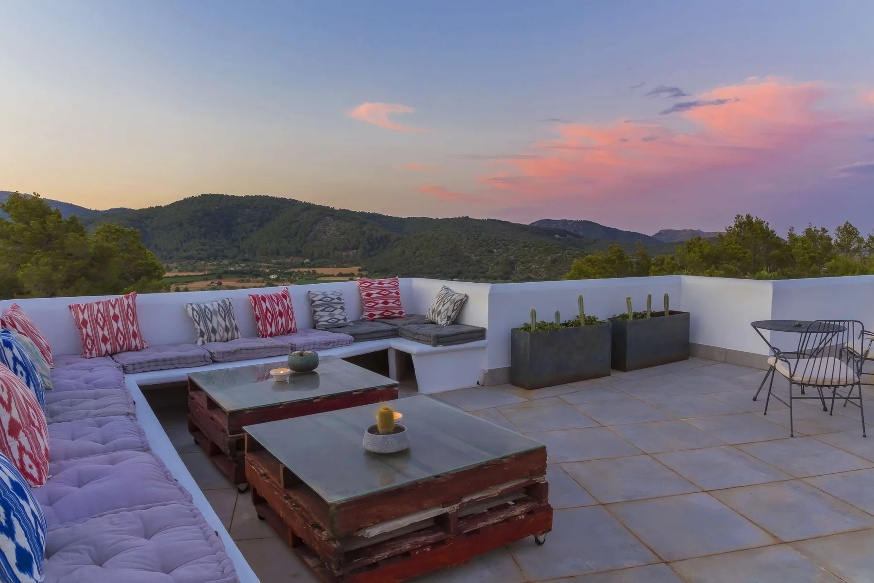 A rooftop terrace with cushioned seating, low tables, and a view of green hills.