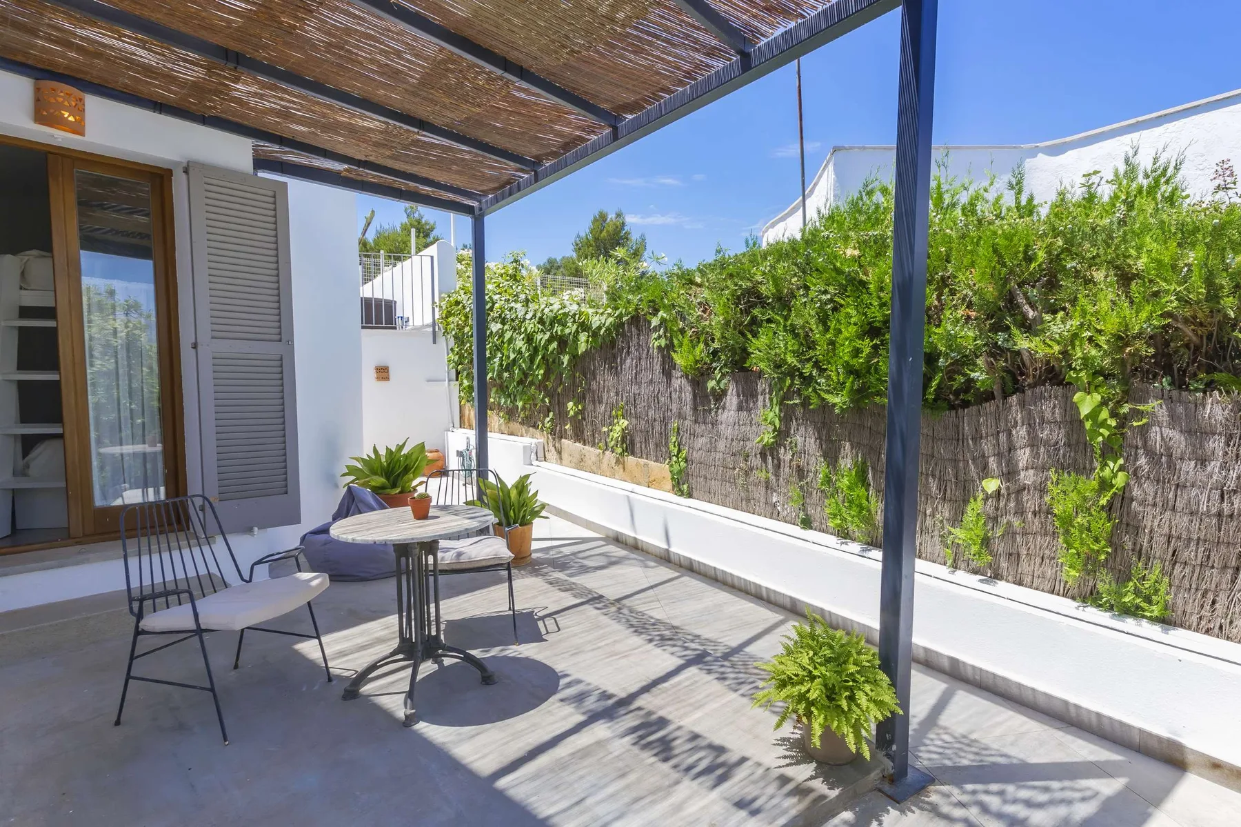 A shaded outdoor patio has seating, a small table, and leafy plants.